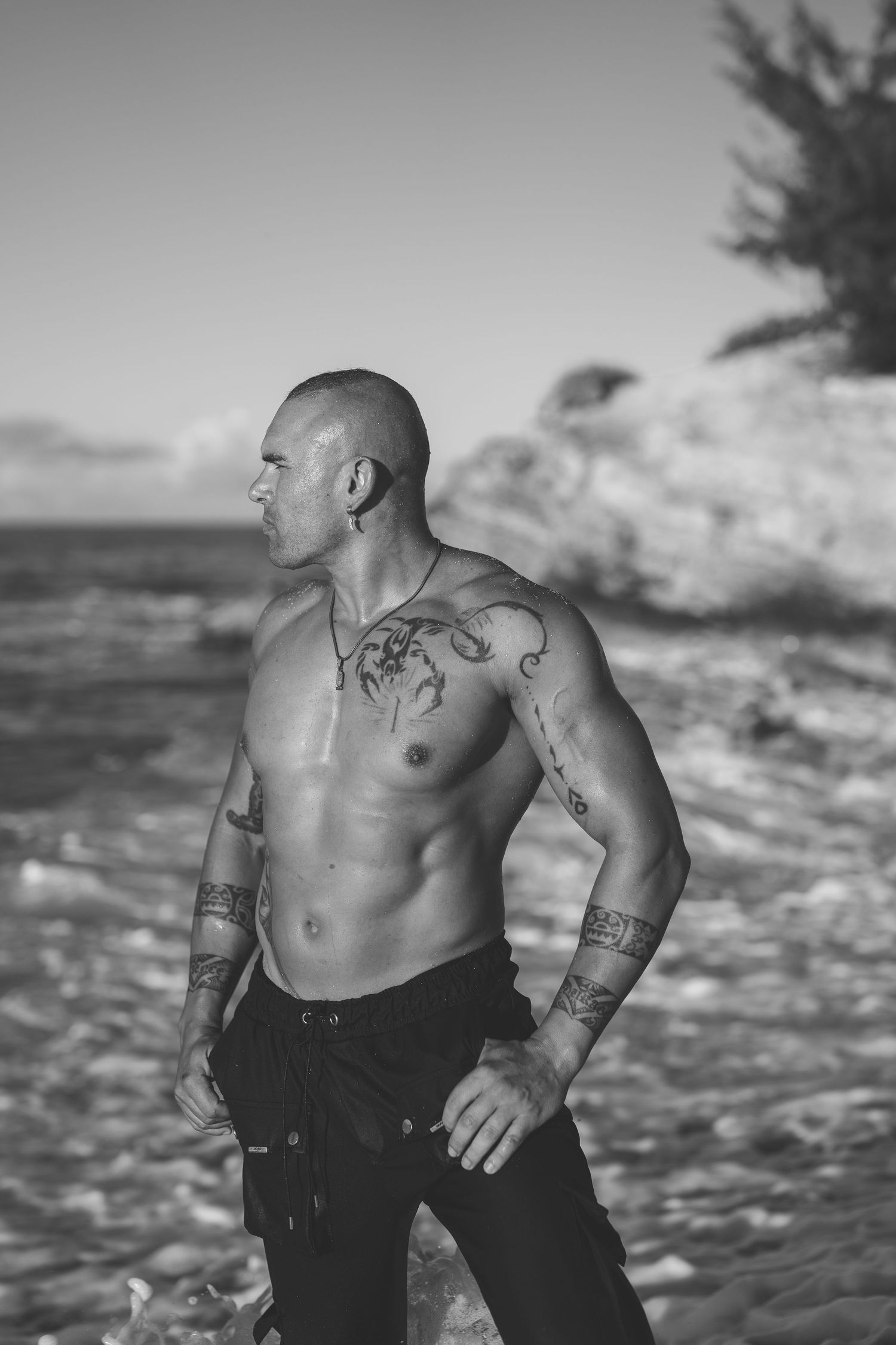 Black and white photo of a muscular tattooed bald man standing shirtless on a rocky beach.