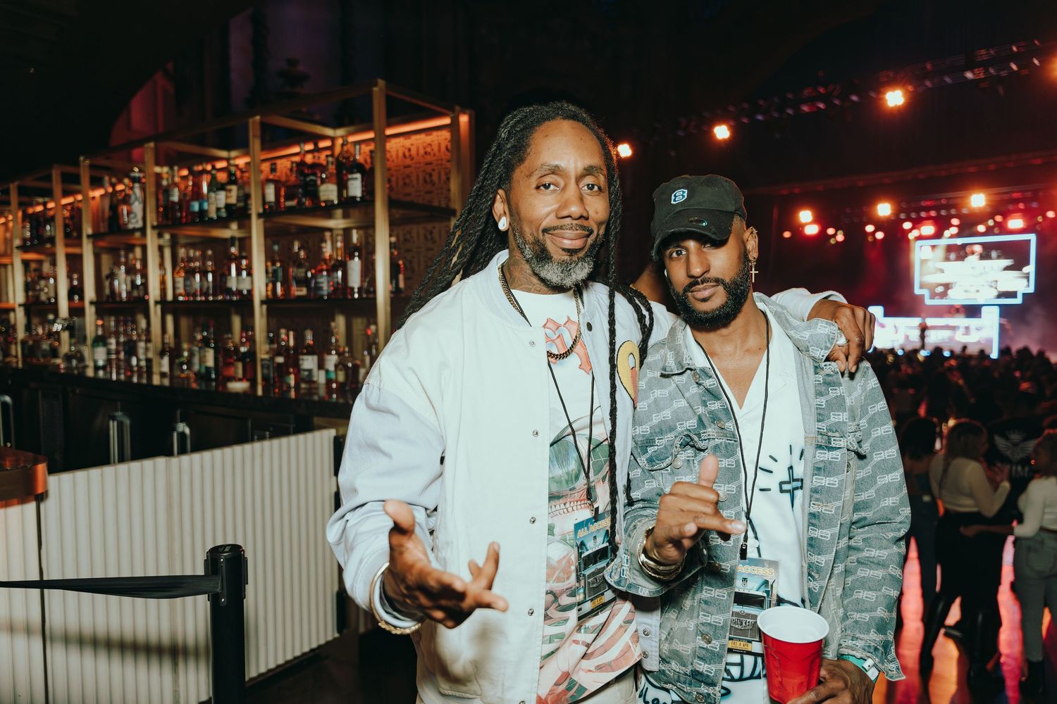 Two men pose together smiling at a bar venue with colorful lights in the background.