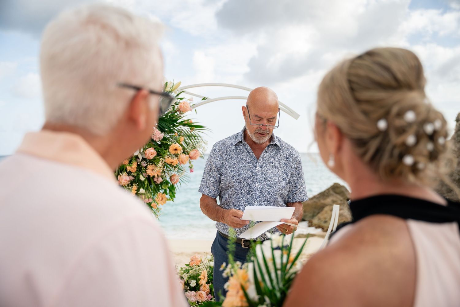 Officiant reading vows to couple during intimate outdoor beach wedding ceremony with floral arch.