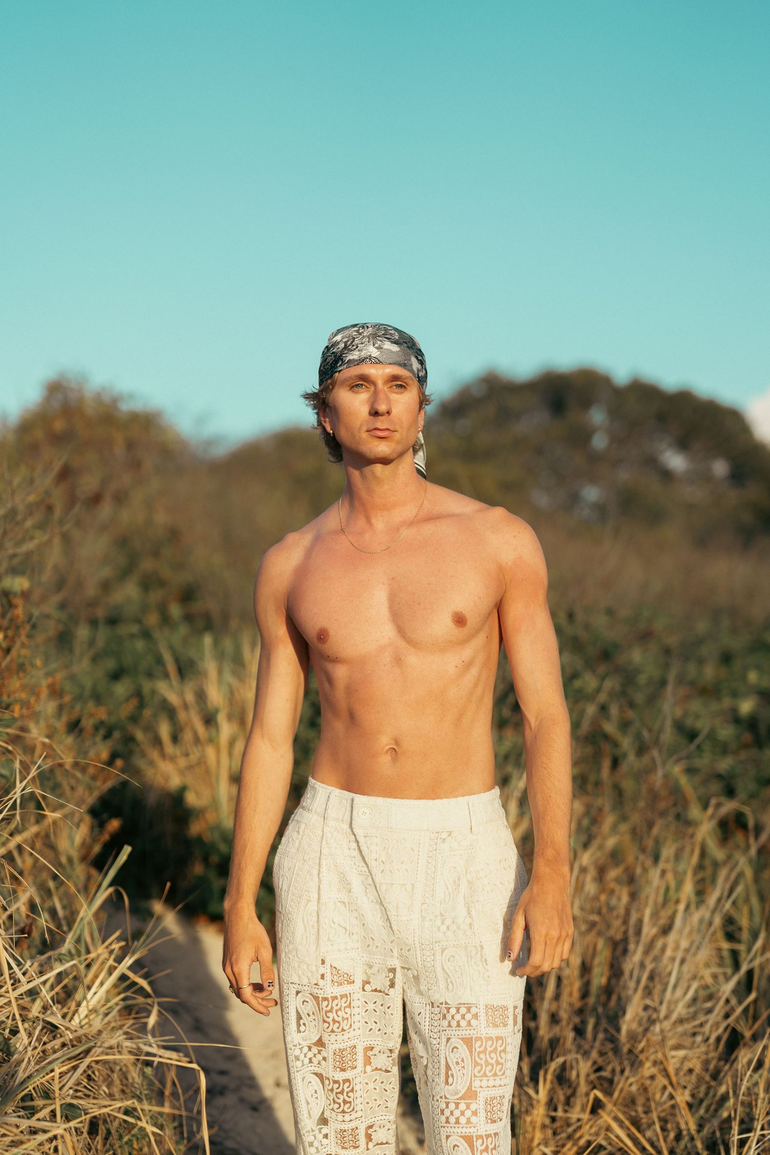 Shirtless man in white lace pants and bandana headband standing in golden hour beach dunes.