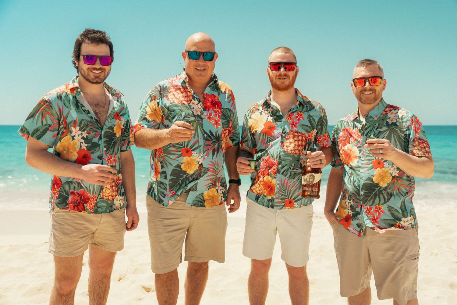 Four men in matching tropical Hawaiian shirts posing on a sunny beach.