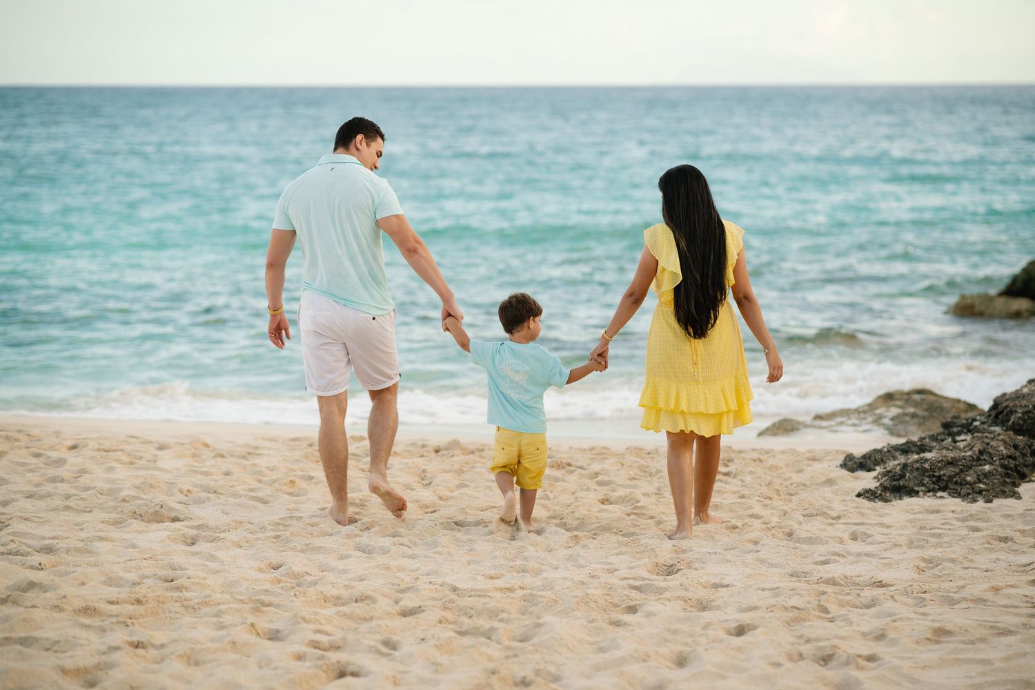 Family of three walking hand in hand toward the ocean on a sandy beach.