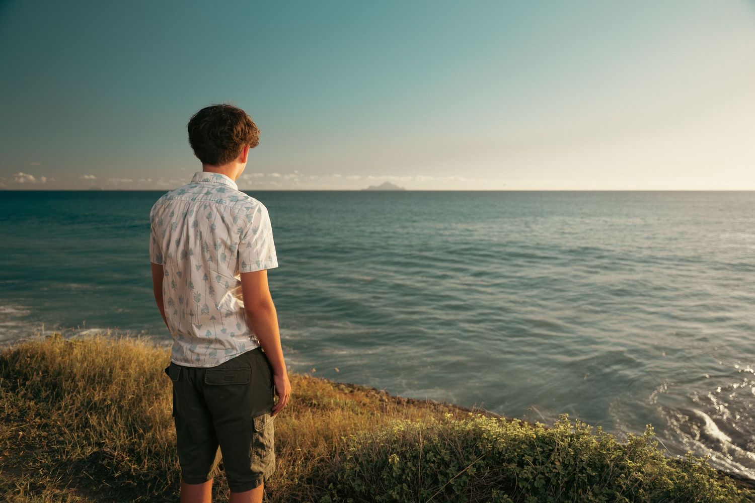Young man in patterned shirt standing on coastal cliff overlooking calm blue ocean at sunset.