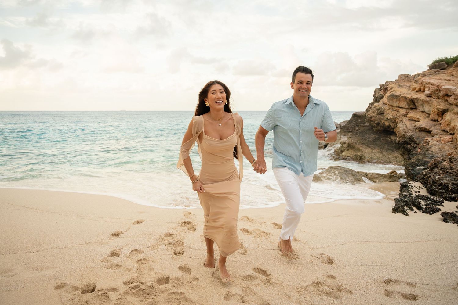 A joyful couple runs barefoot along a sandy beach near rocky cliffs, woman in beige dress.