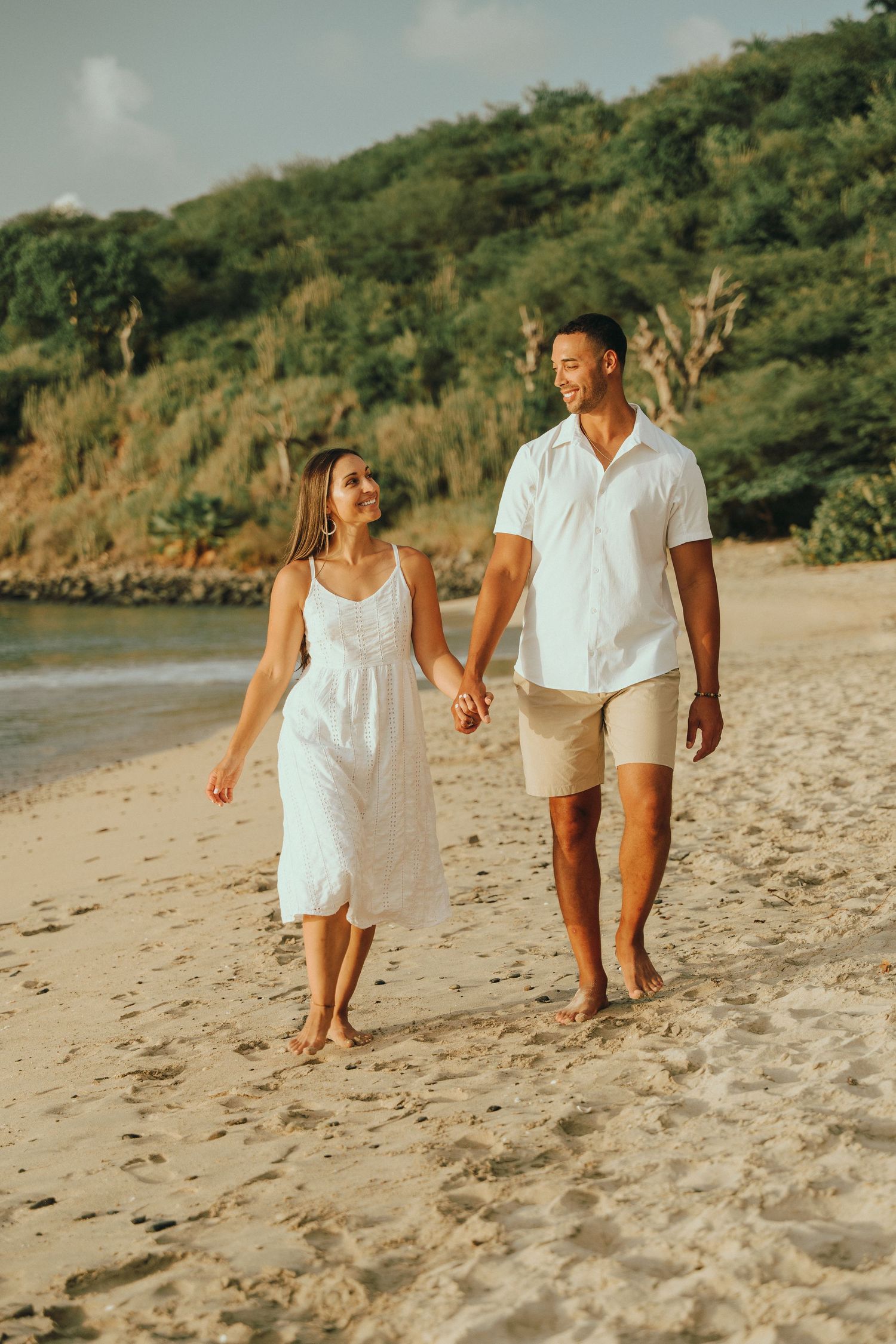 Happy couple walking hand-in-hand barefoot along a sandy tropical beach, woman in white dress and man in white shirt.