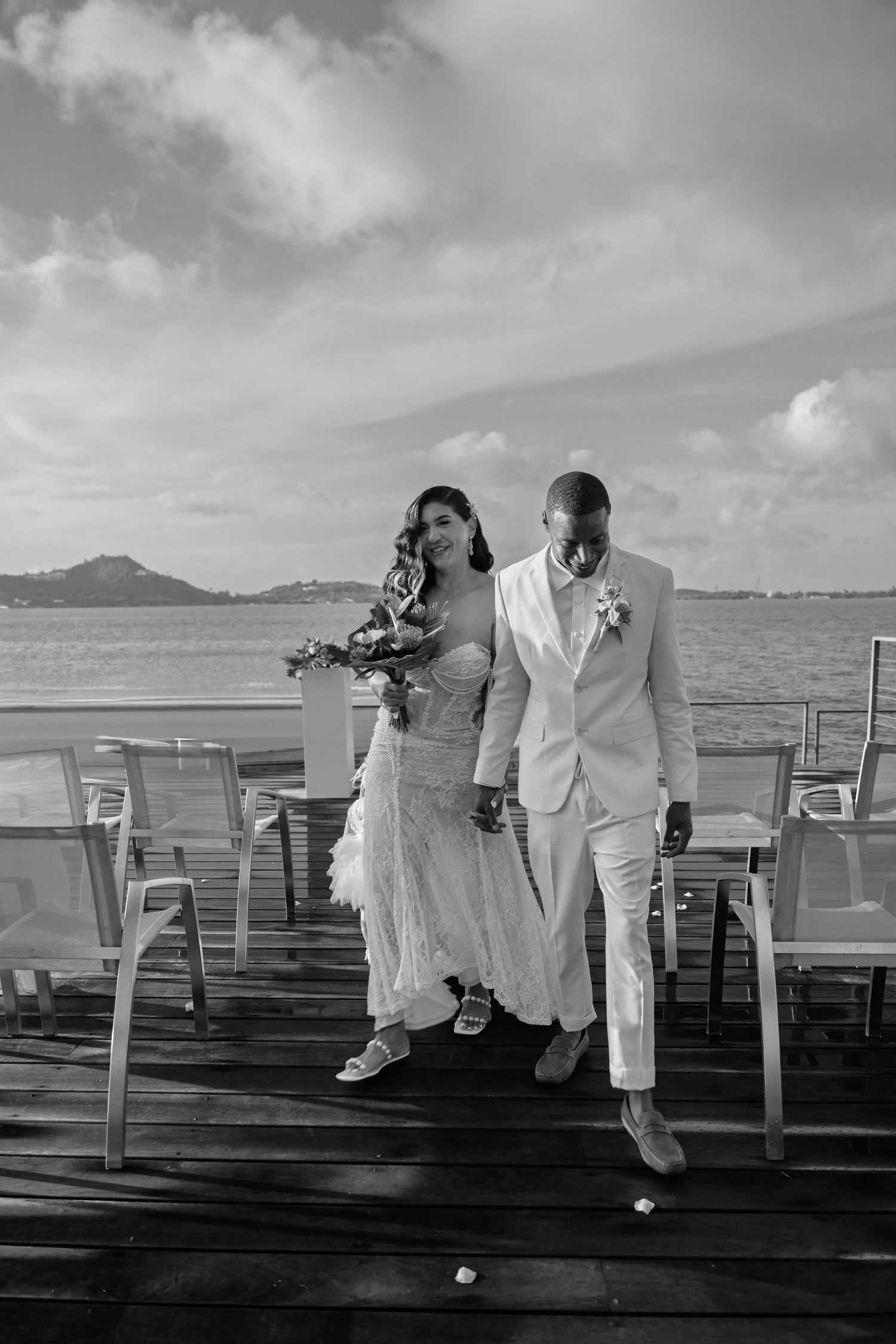 Black and white photo of newlywed couple walking on waterfront deck after wedding ceremony.