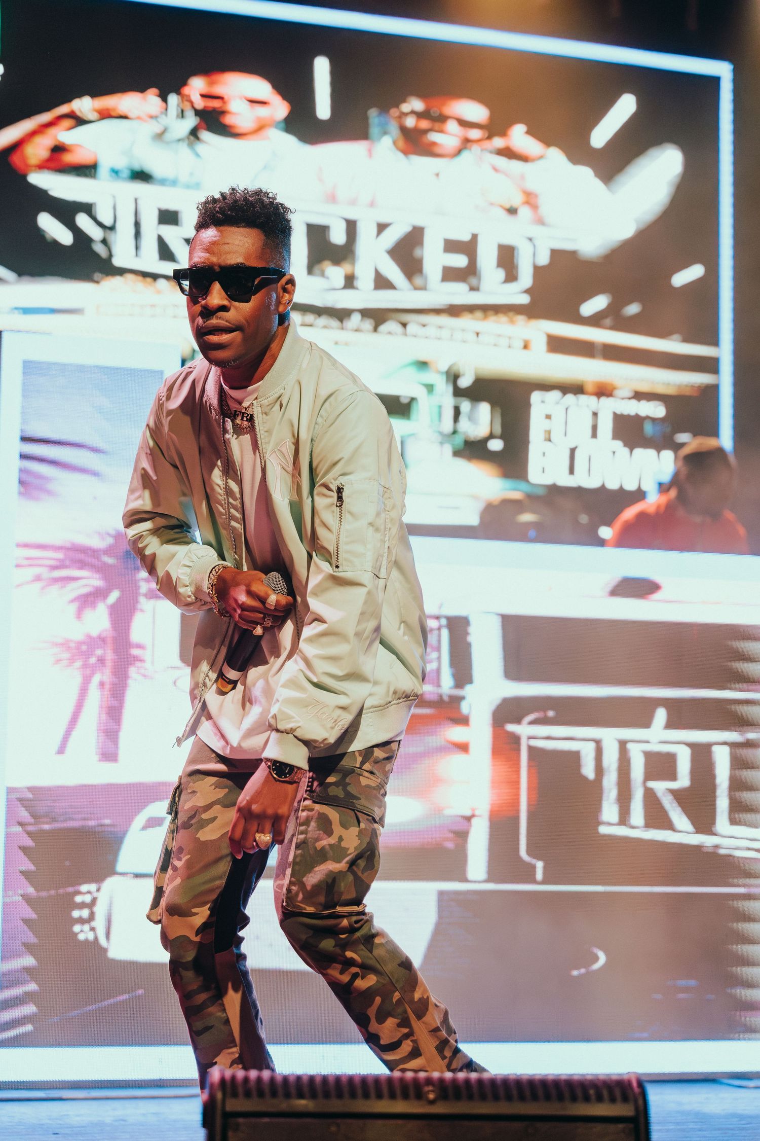 Male artiste in camo pants and sunglasses performing energetically on a lit concert stage.