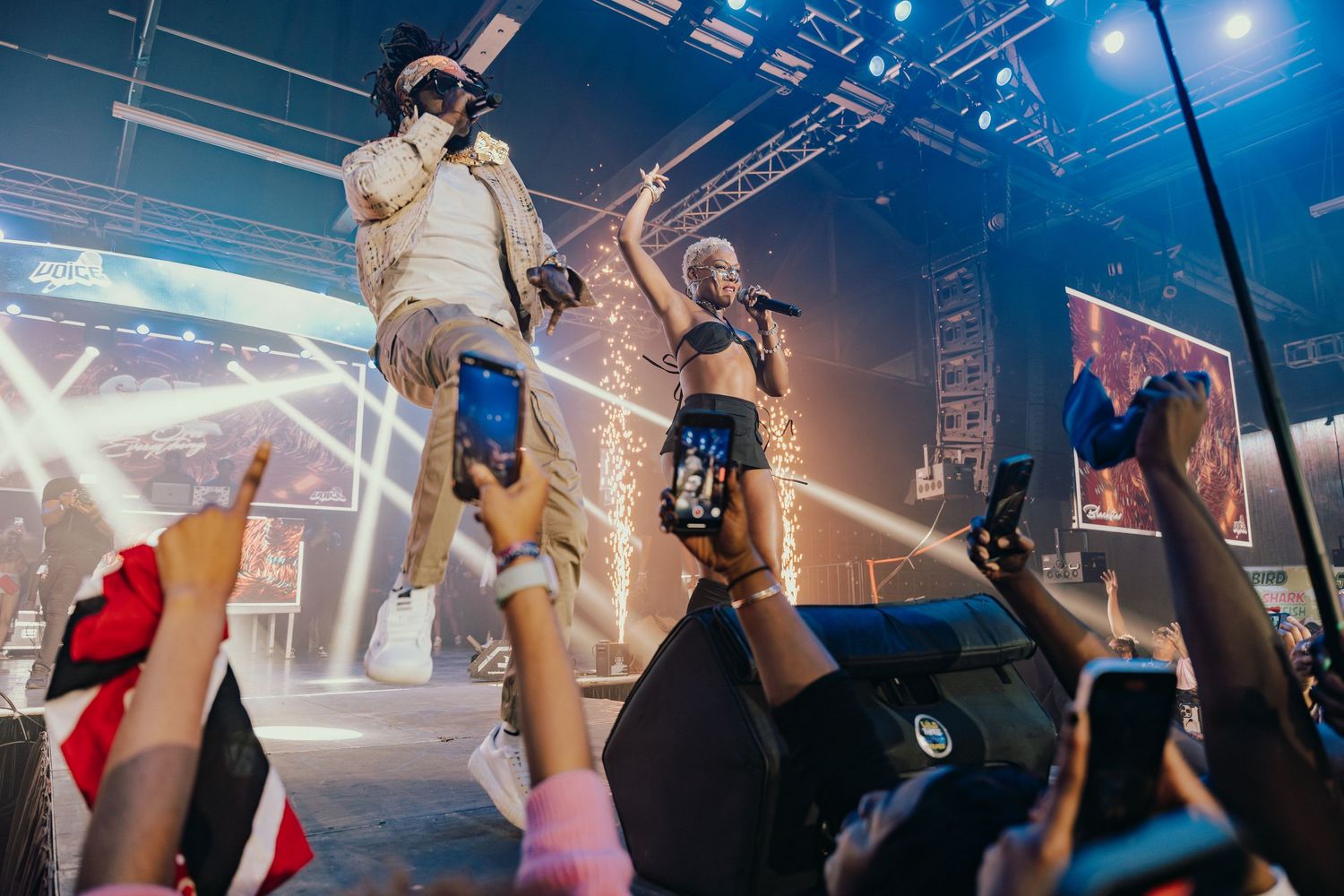 Two performers on stage engaging with crowd holding up phones at a live concert event.