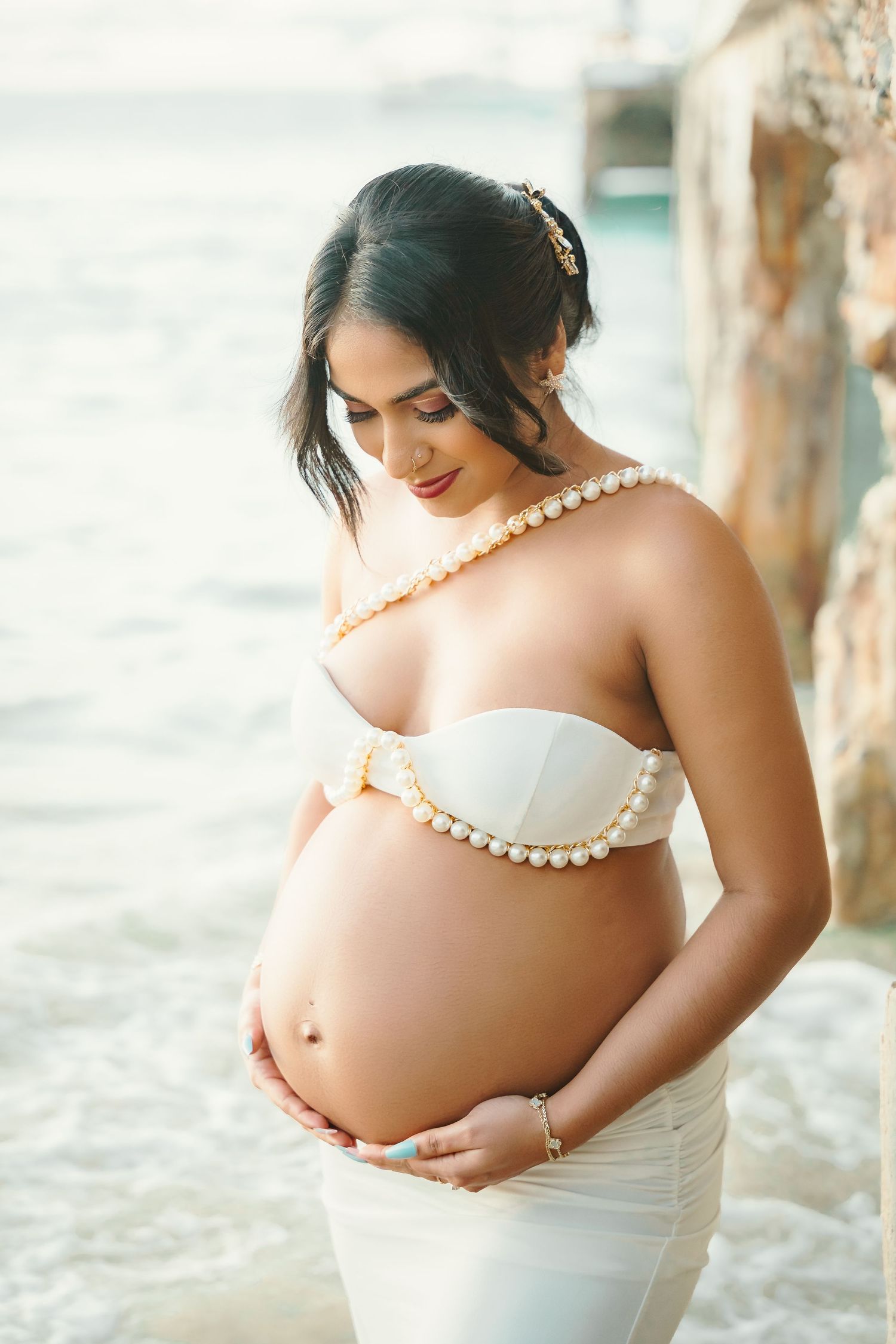 Beautiful pregnant woman in white pearl bikini and skirt cradles belly on tropical beach.