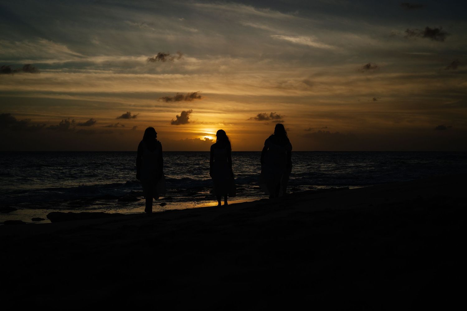 Three silhouetted figures stand together watching a dramatic ocean sunset.