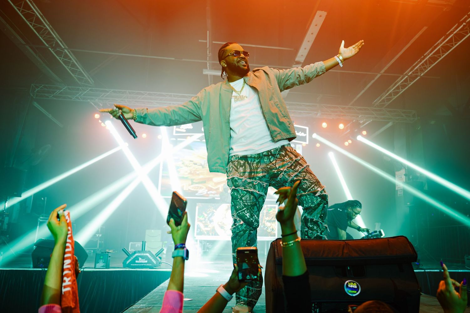 Male performer spreads arms wide on stage while crowd reaches up under teal concert lighting.