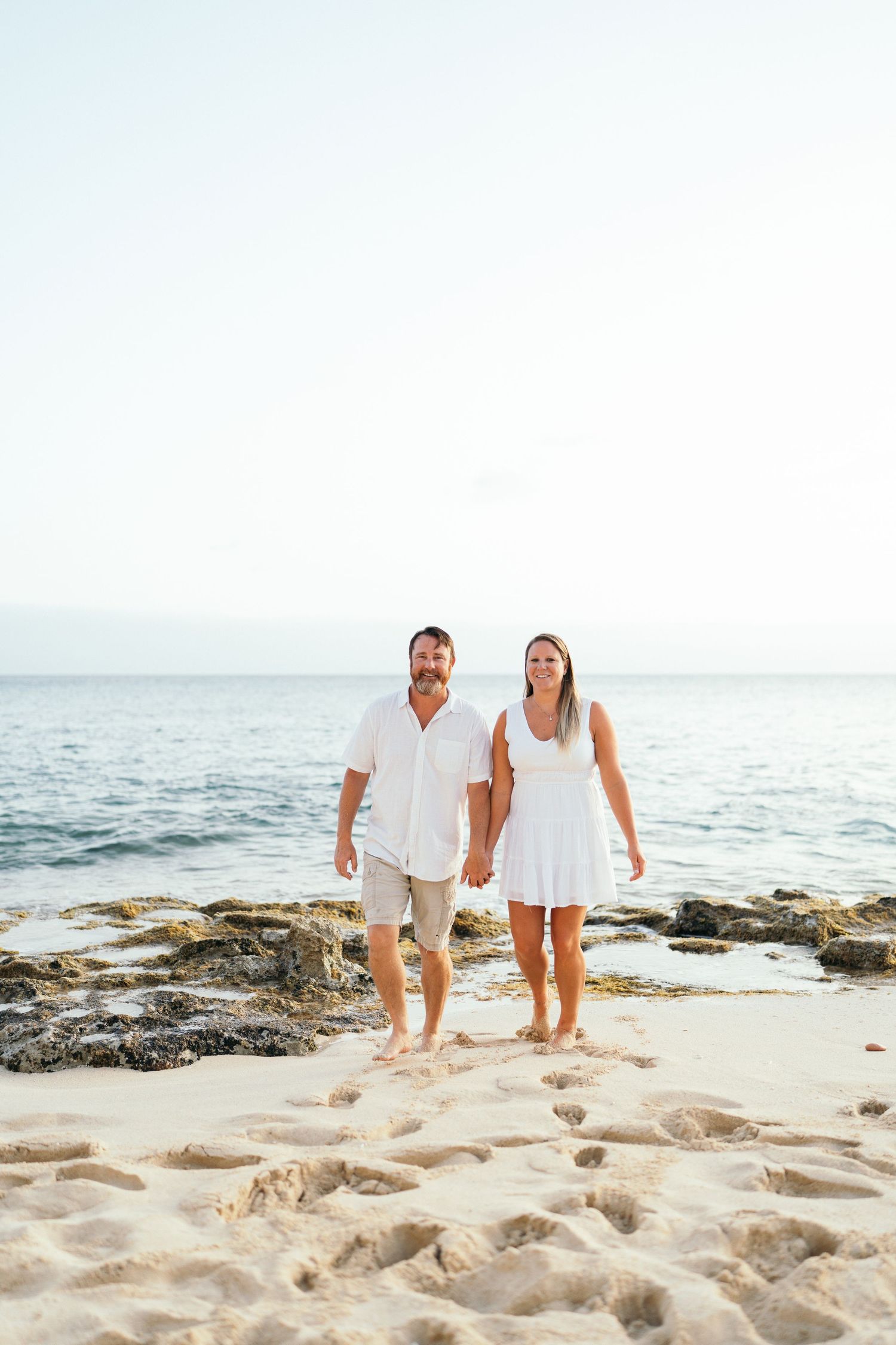 A couple in white outfits holds hands while walking on a sandy beach with turquoise ocean behind them.