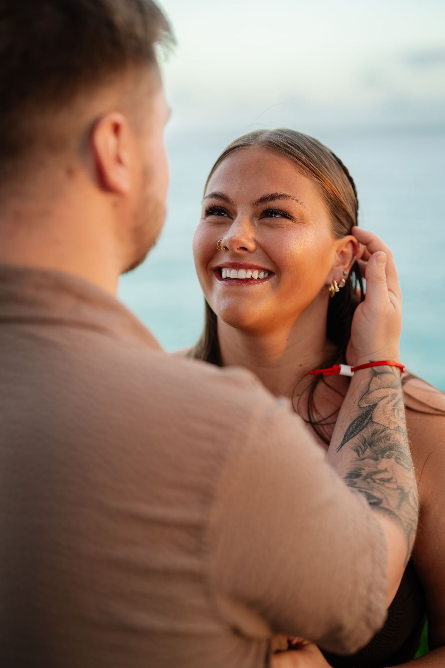 Joyful woman smiling as tattooed partner gently tucks hair behind her ear by turquoise ocean.