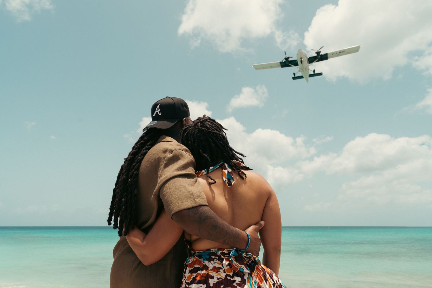 Couple embracing on a tropical beach watching a small plane fly overhead against a blue sky.