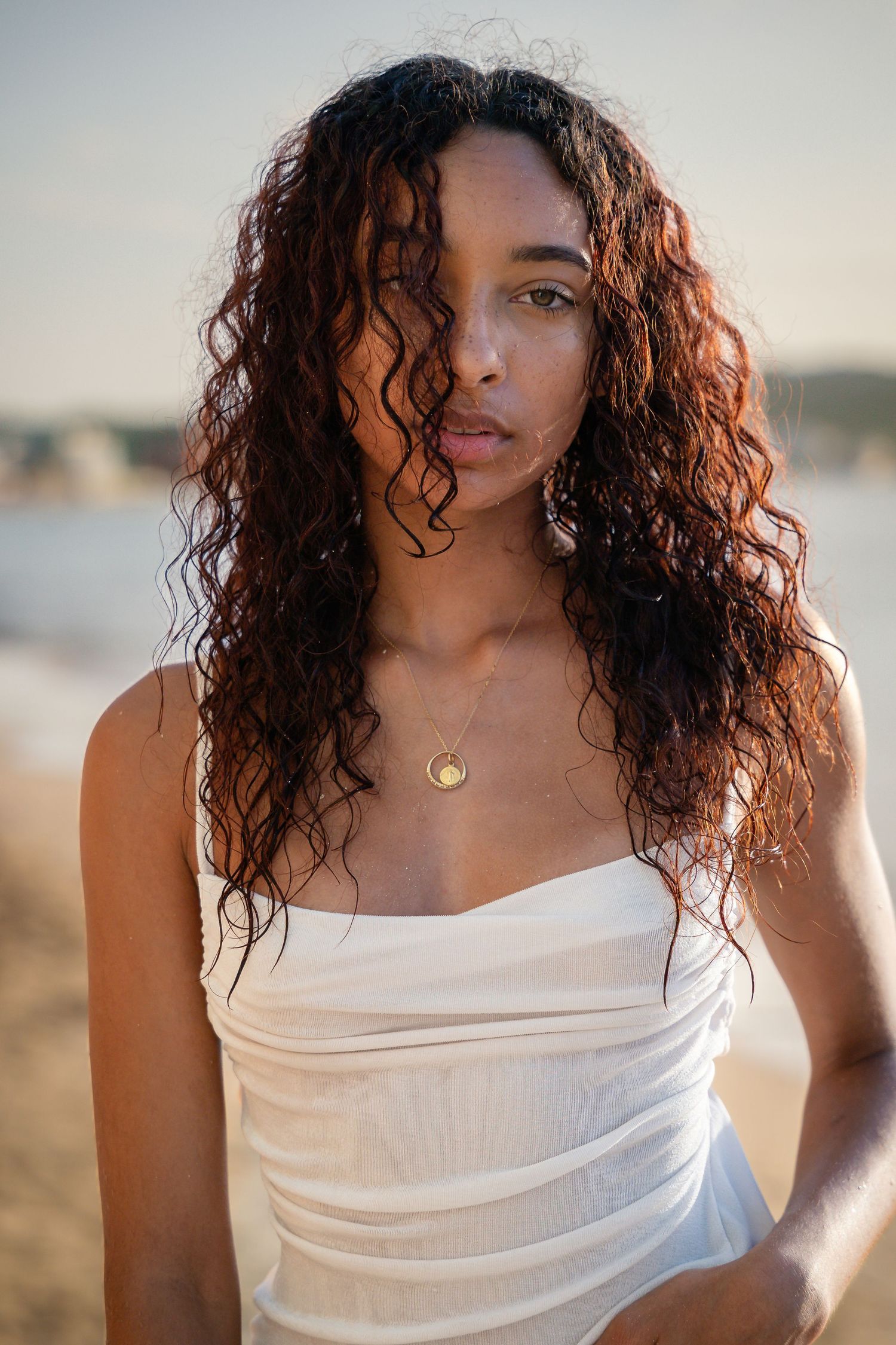 Young woman with long curly auburn hair wearing a white strapless top and gold necklace at a sunny beach.