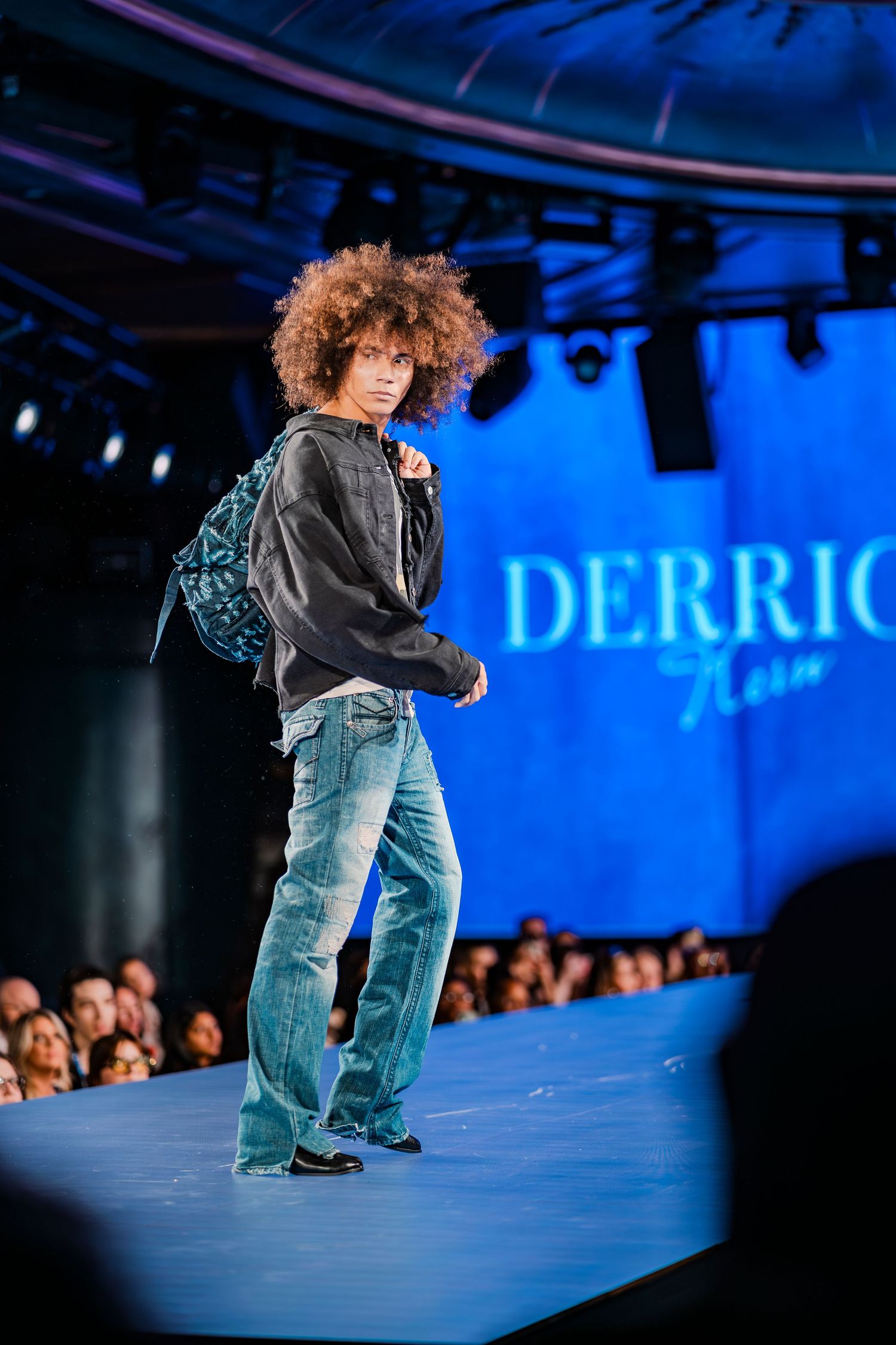 Model walks runway in baggy jeans and dark hoodie with patterned backpack at Derrick fashion show.