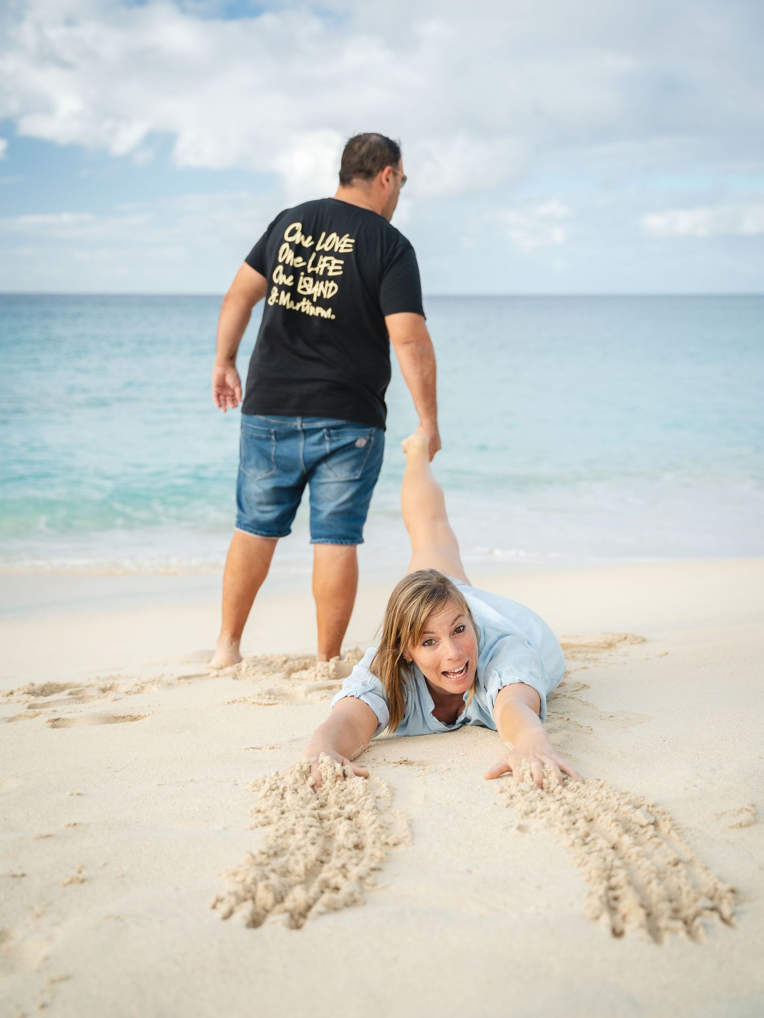 Playful couple on a tropical beach, woman lying in the sand laughing while man stands behind her near the ocean.