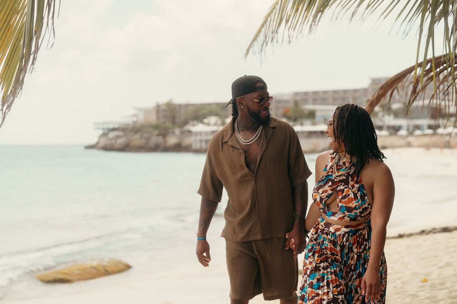 A couple walks hand-in-hand on a tropical beach, man in brown outfit and woman in colorful dress.