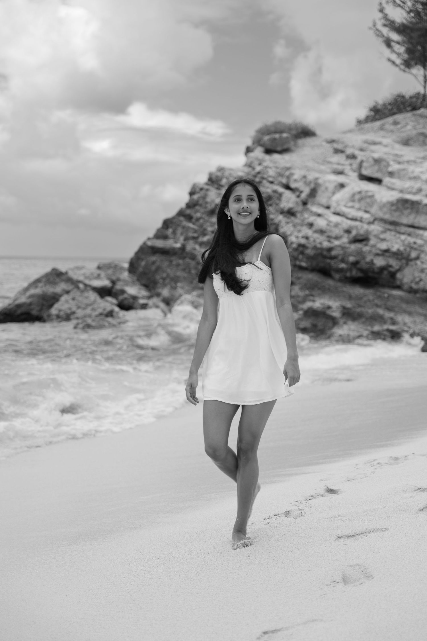 Black and white photo of a young woman in a white dress walking barefoot on a sandy tropical beach.