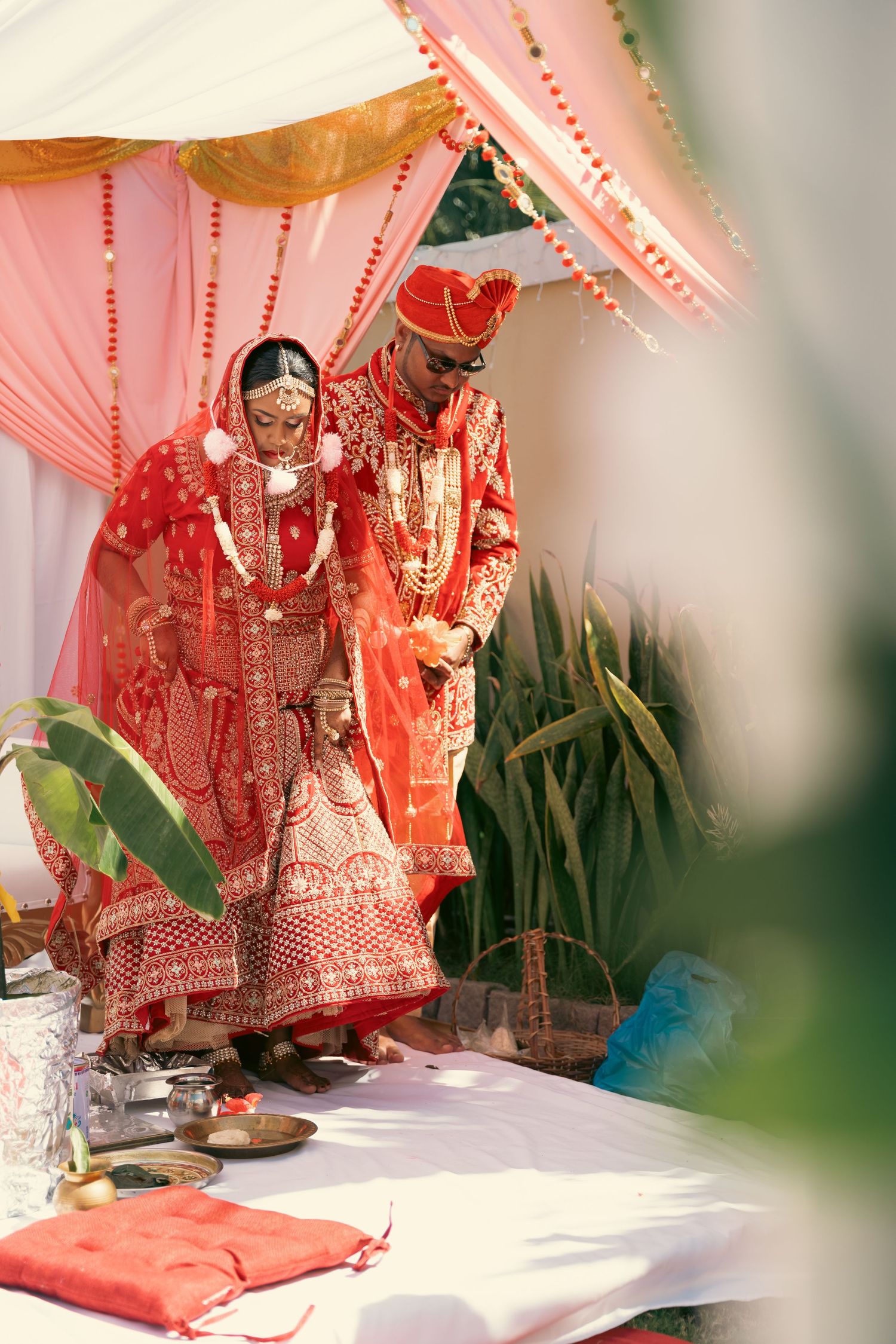 Indian bride and groom in matching red attire performing traditional wedding rituals under mandap.