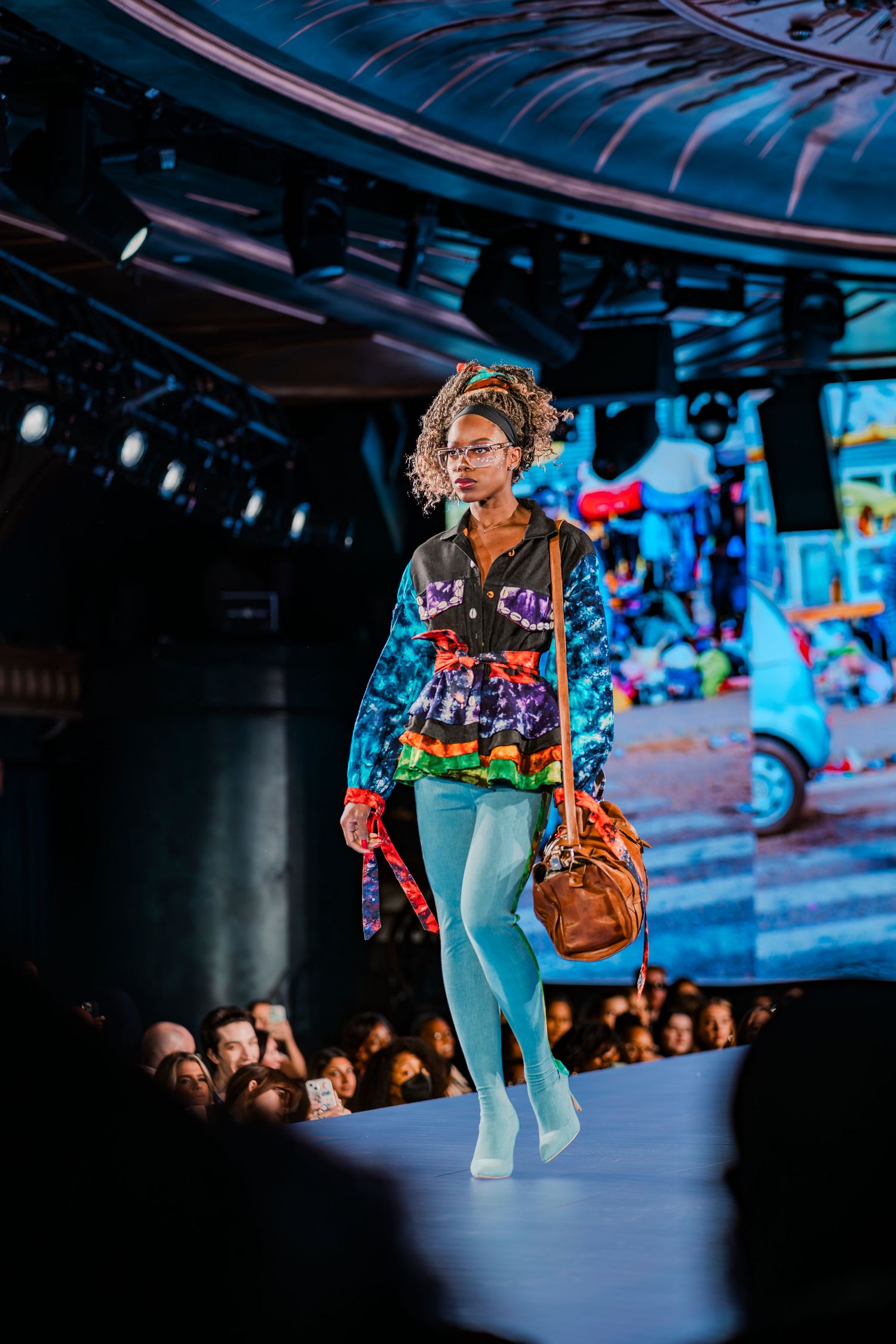 Model walks runway in colorful eclectic outfit with teal tights, patterned jacket, and brown bag at fashion show.