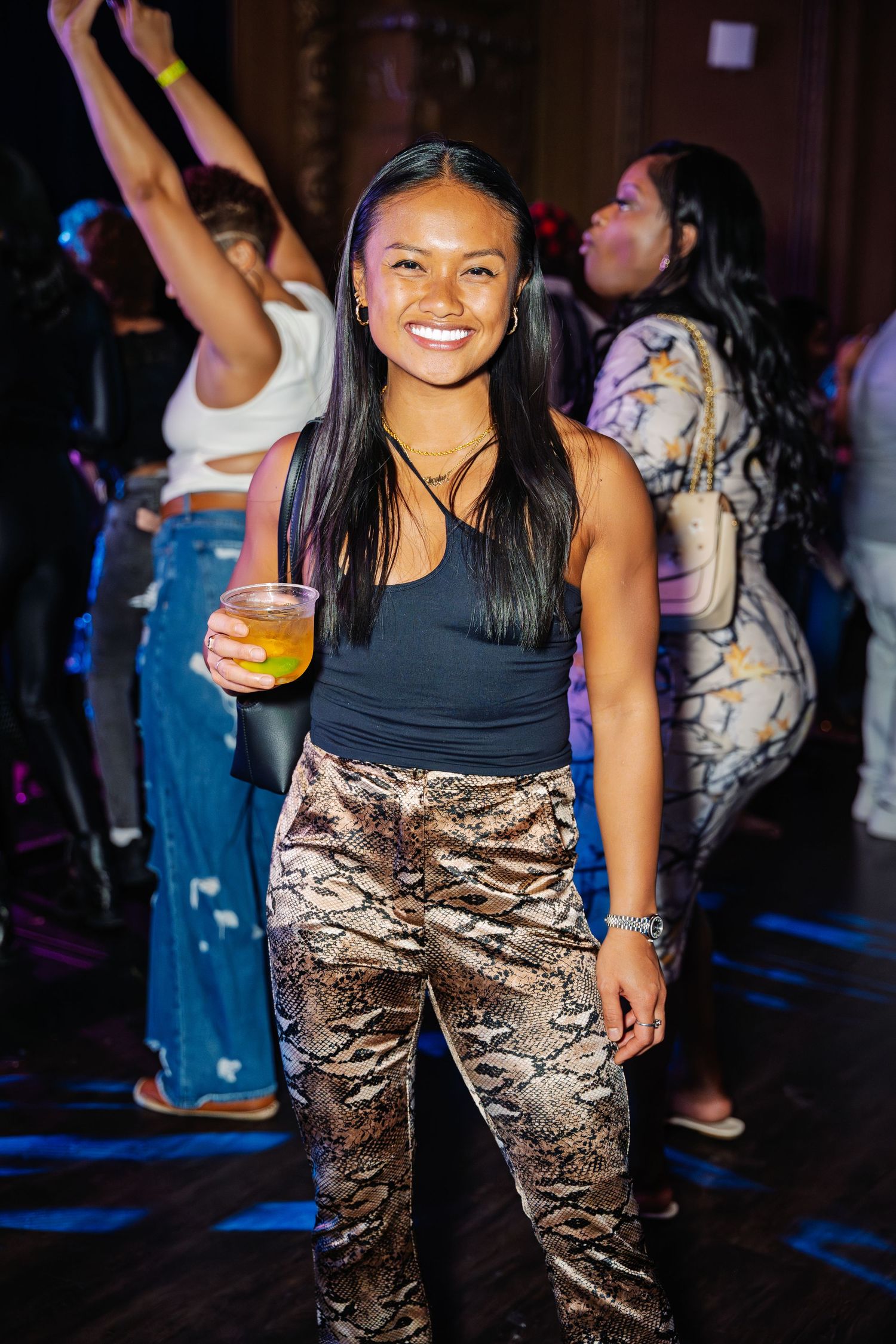 Smiling woman in snakeskin pants holds a drink on a lively dance floor.