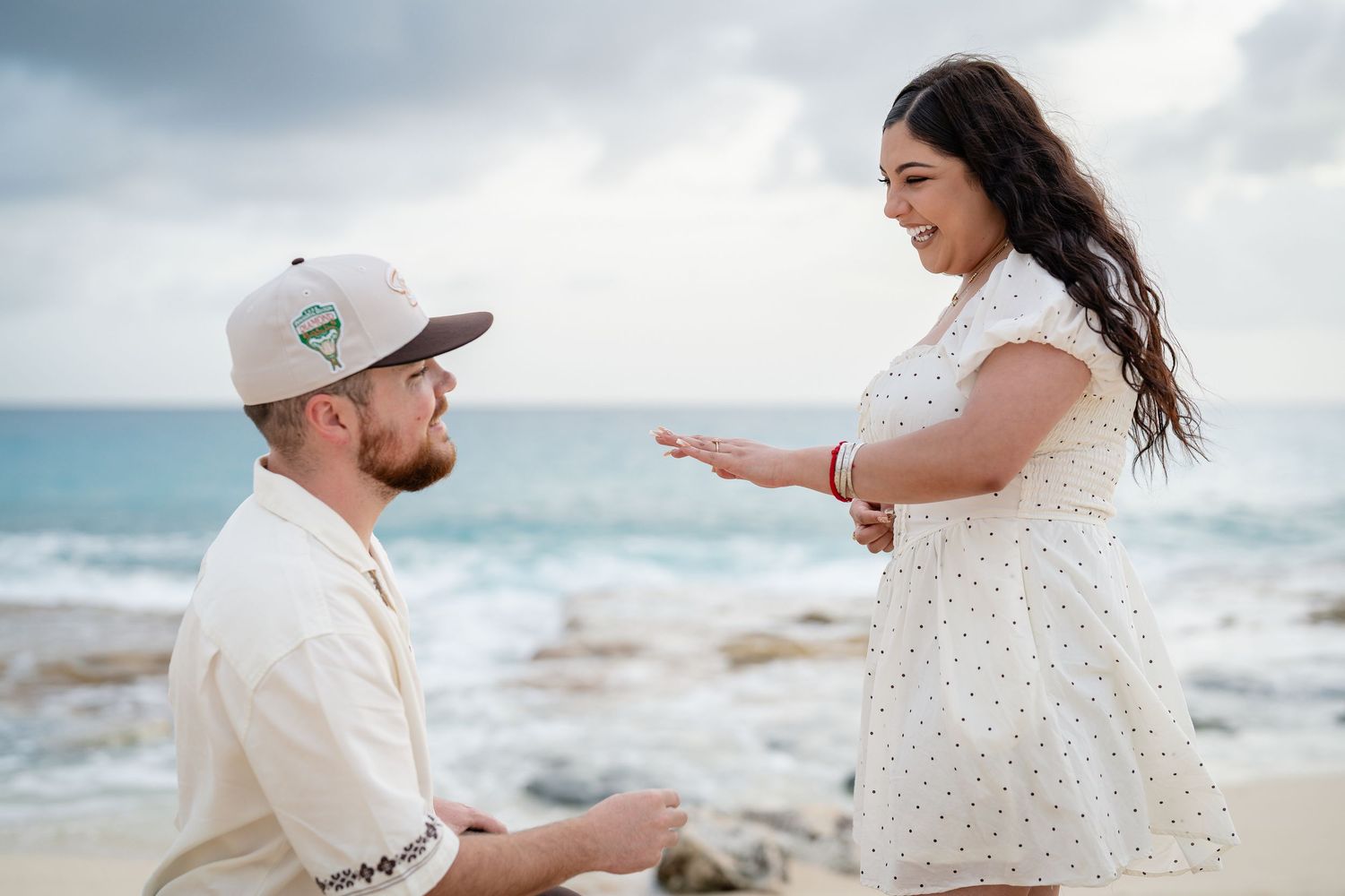 Man on one knee proposing to surprised woman on rocky beach with ocean backdrop.
