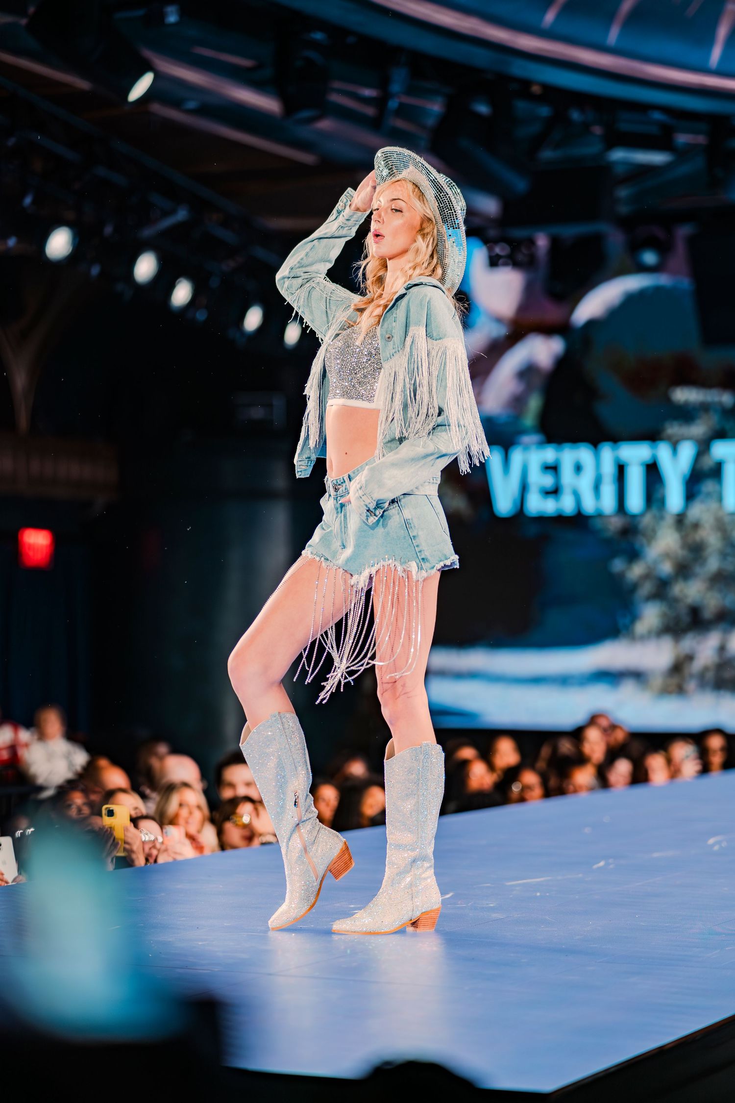 Model walks runway in light blue fringe cowgirl outfit with matching hat and white boots at fashion show.