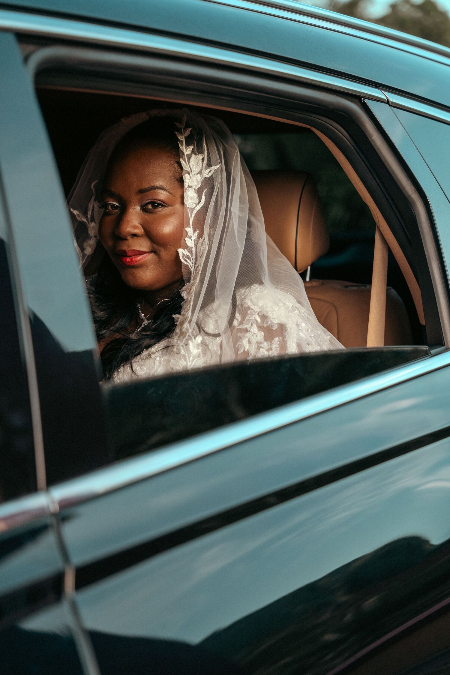 Smiling Black bride in white lace gown and veil seated in luxury car on wedding day.