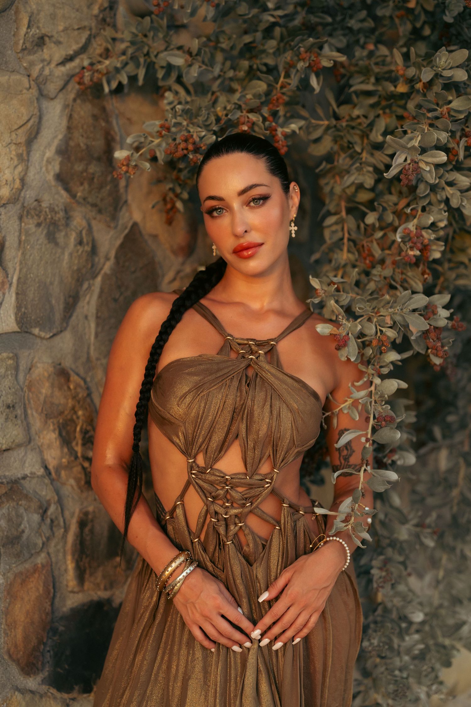 Woman with a long braid in a gold macrame cutout dress posing against a stone wall with berry plants.