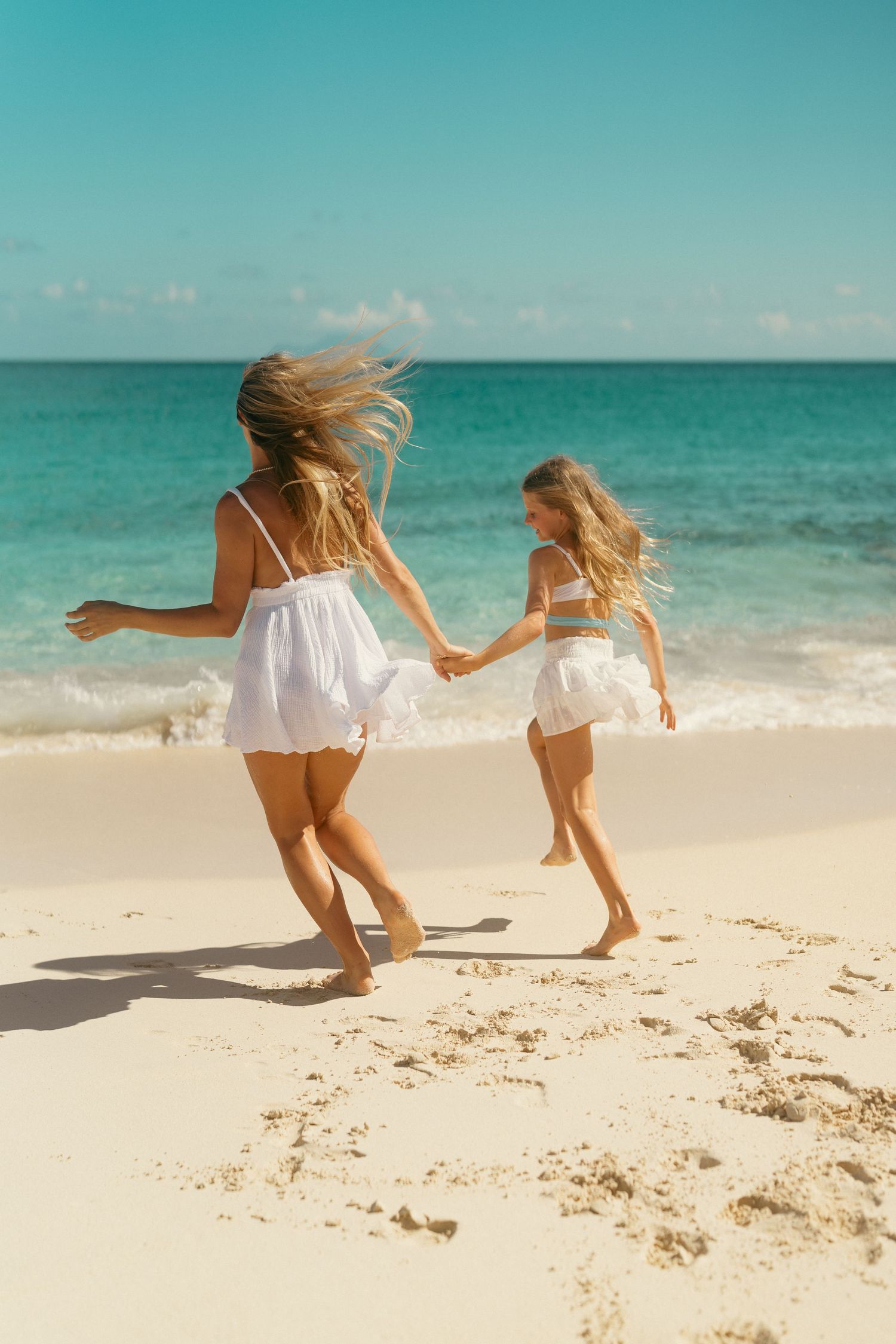Two girls in white dresses run hand in hand along a tropical sandy beach.