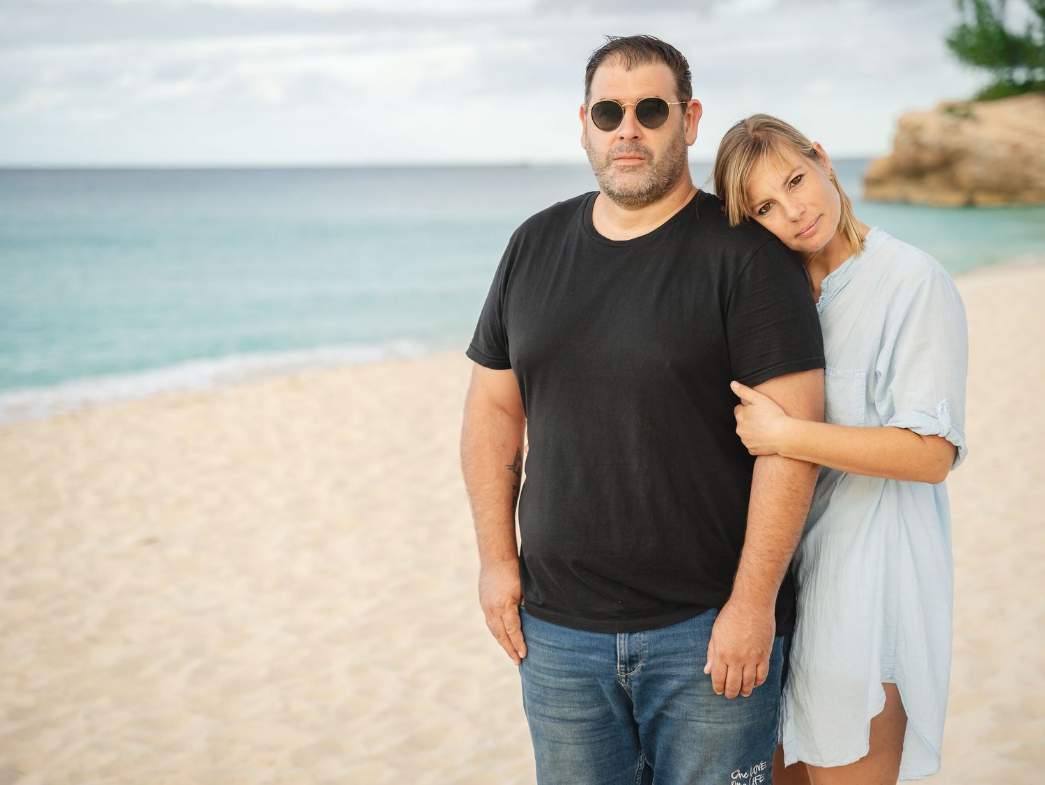 Couple posing together on a tropical beach, man in black t-shirt and woman in blue dress with turquoise ocean behind.