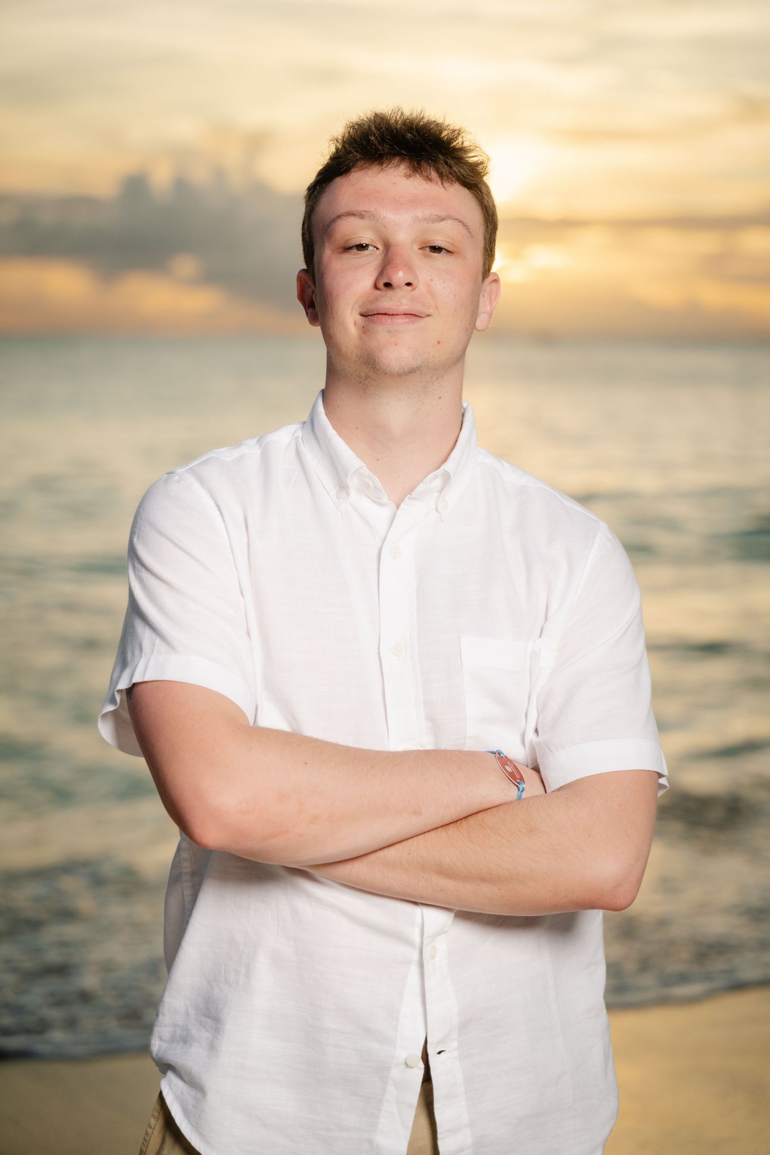 Young man in white button-up shirt standing with arms crossed on a beach at golden sunset.