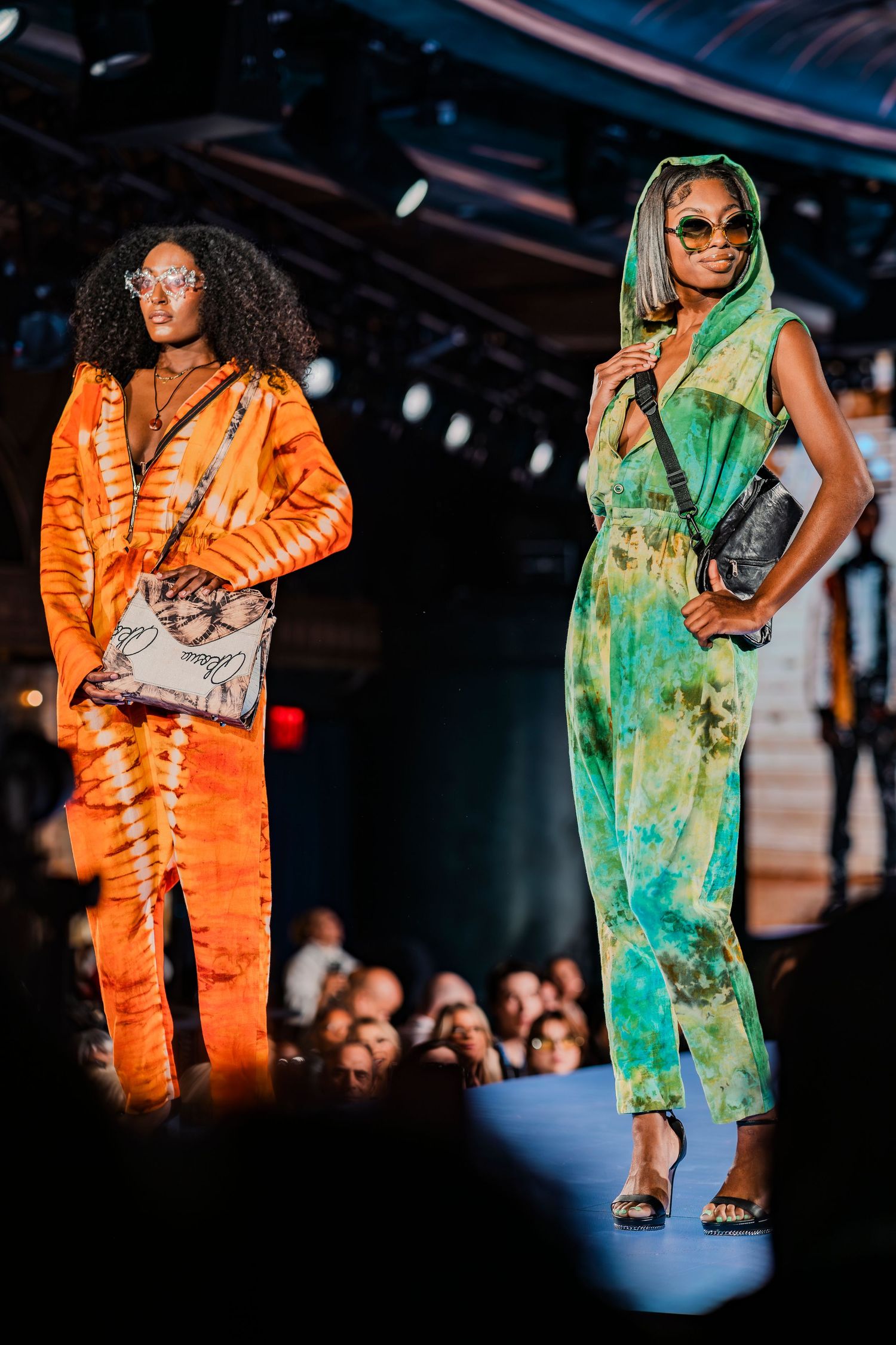 Two models on runway wearing vibrant tie-dye jumpsuits in orange and green, accessorized with crossbody bags and sunglasses.