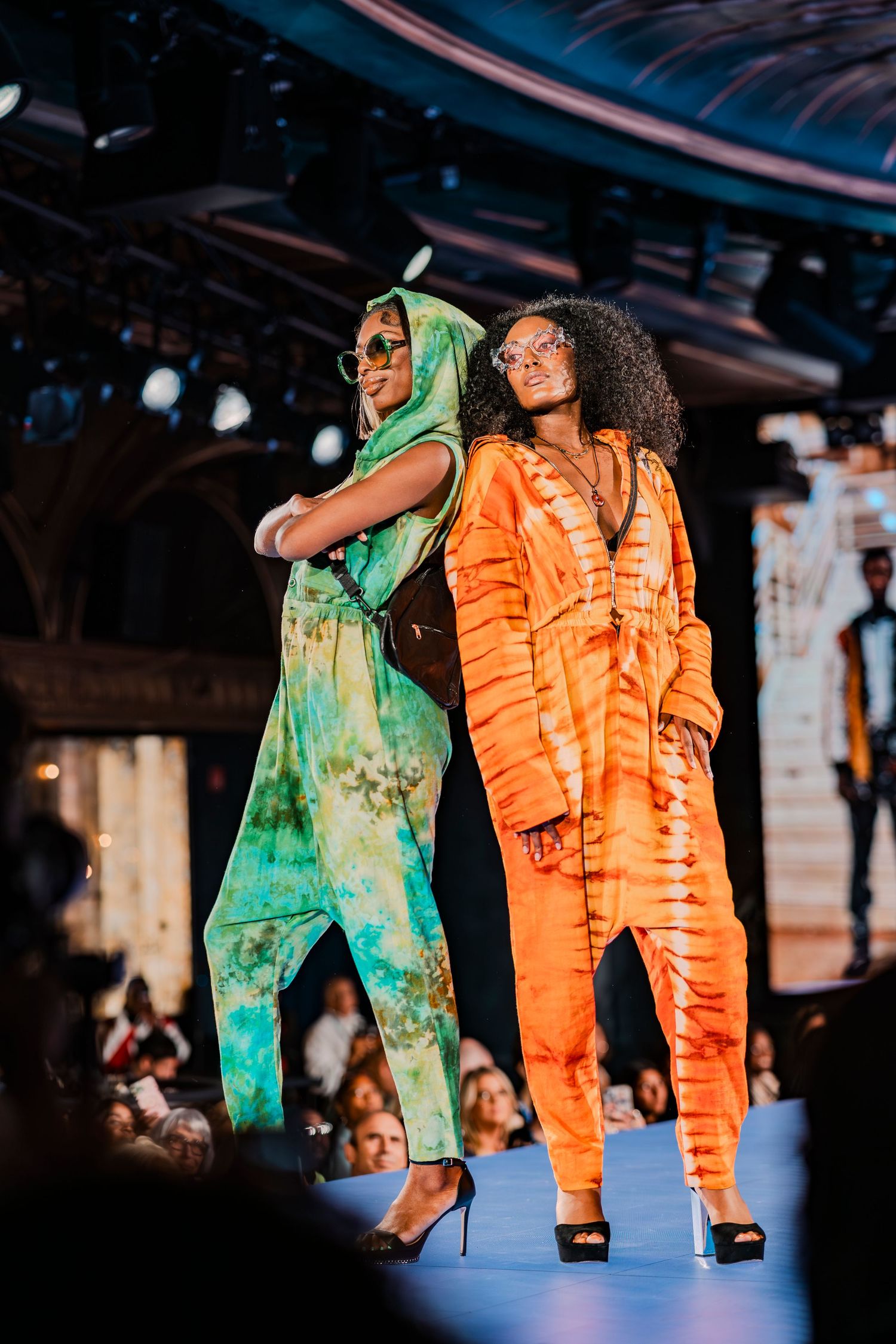 Two models walk the runway in vibrant tie-dye jumpsuits, one green and one orange, at a fashion show.