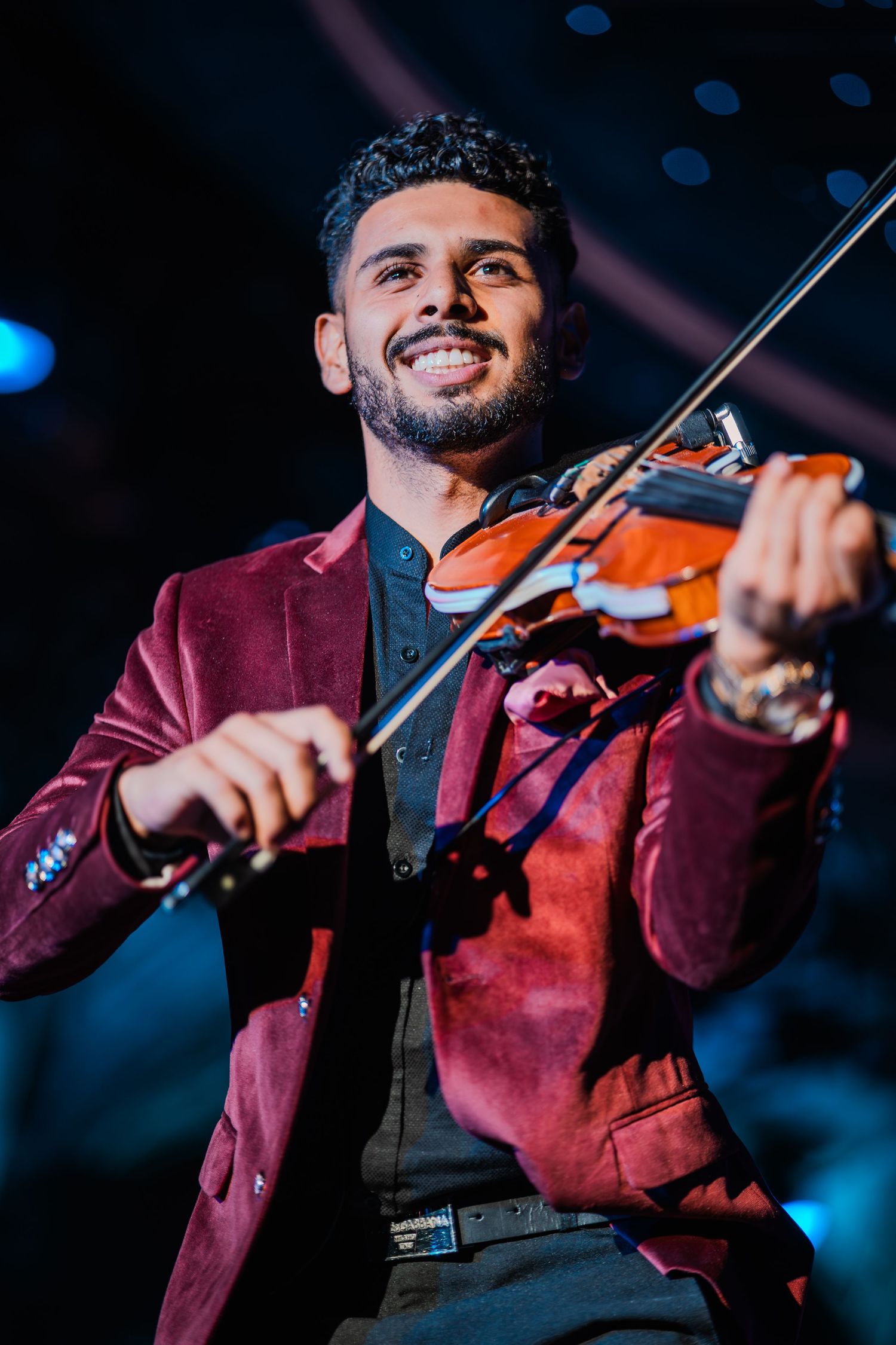 Smiling male violinist performing on stage in a burgundy velvet blazer, playing violin under dramatic blue stage lighting.