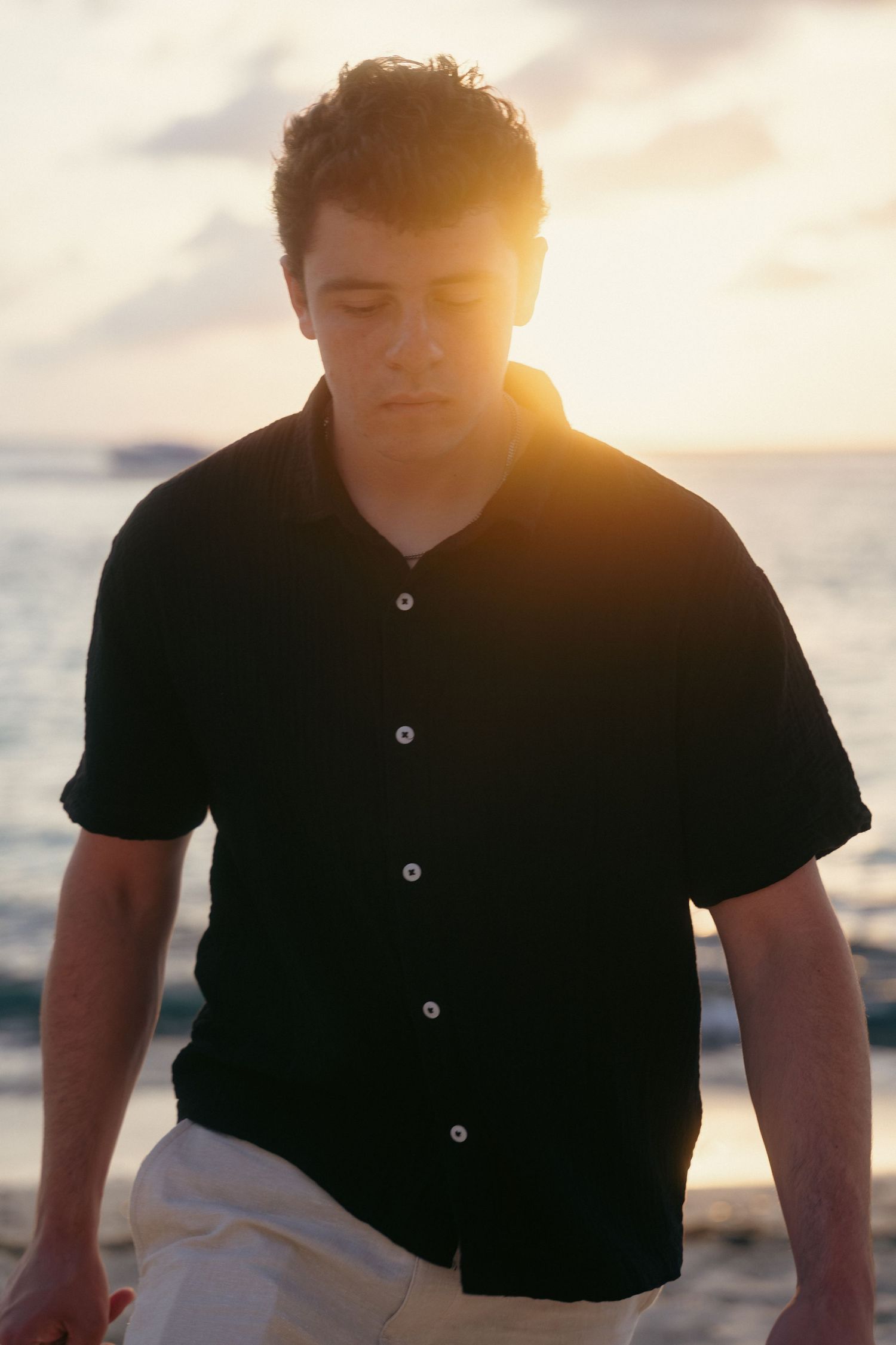 Young man in black polo shirt stands on beach at golden sunset with ocean glowing behind him.