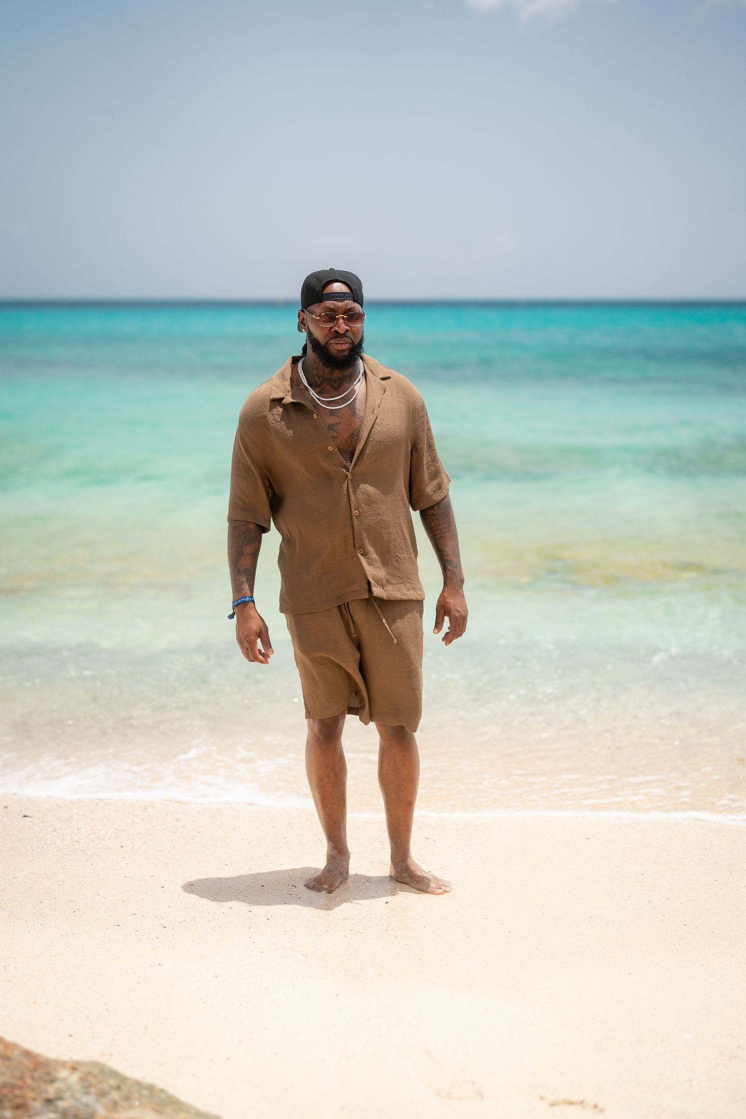 Tattooed man in tan shorts set and cap stands barefoot on white sand beach with turquoise water behind him.