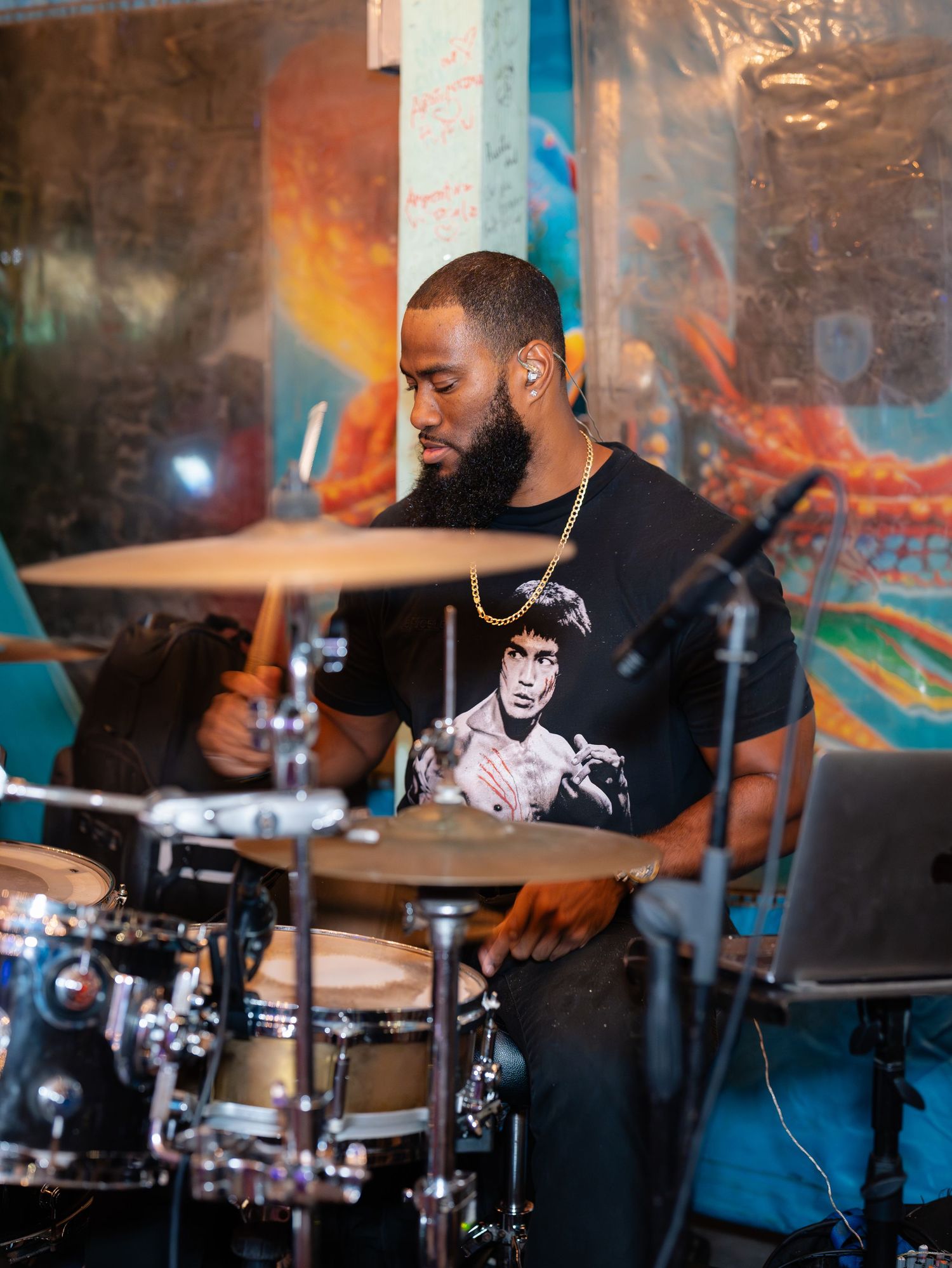 Bearded Black male drummer performing live music wearing a Bruce Lee graphic tee and gold chain.