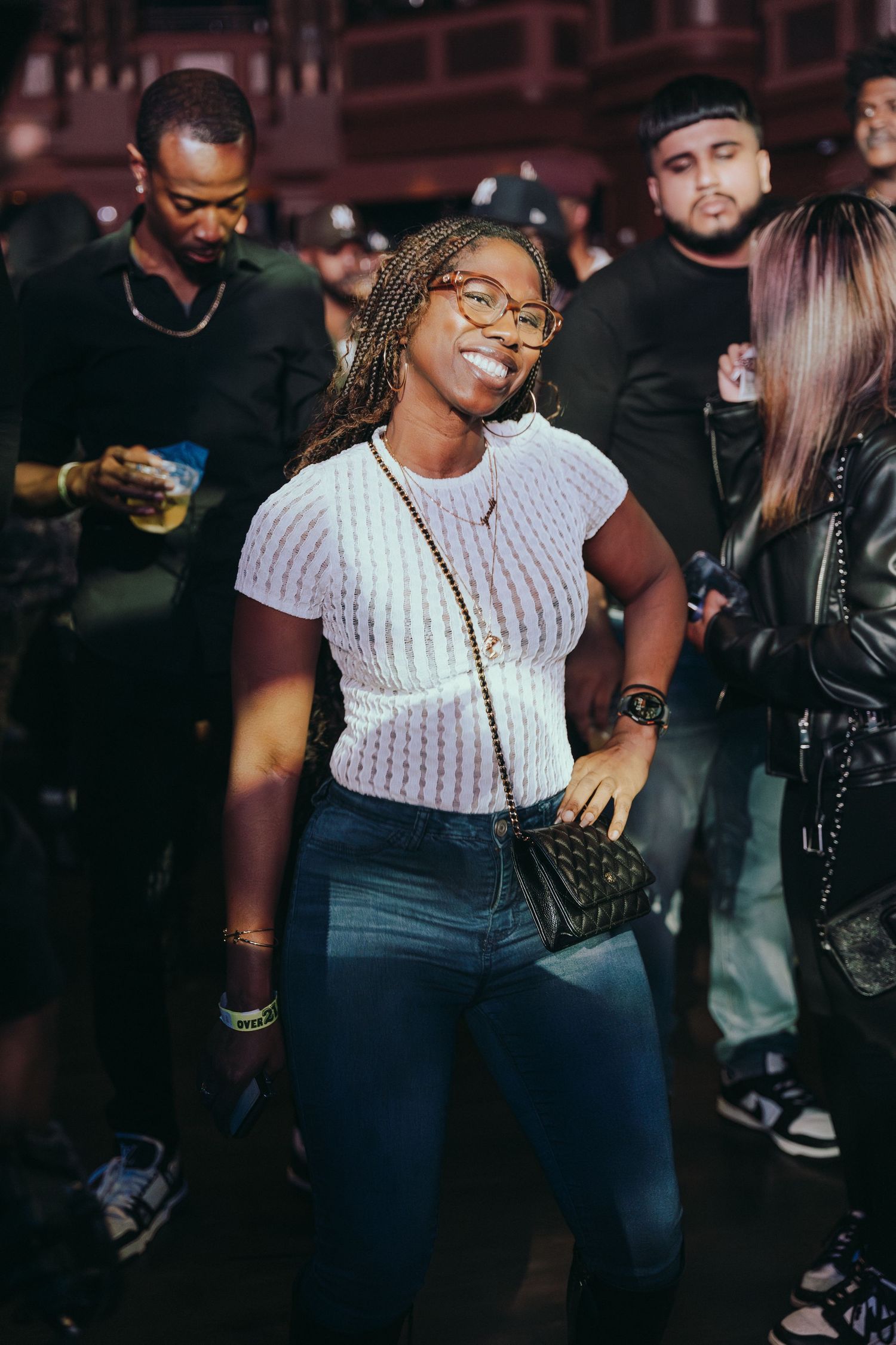 Young woman with braids and glasses smiles while dancing at a crowded nightclub event.