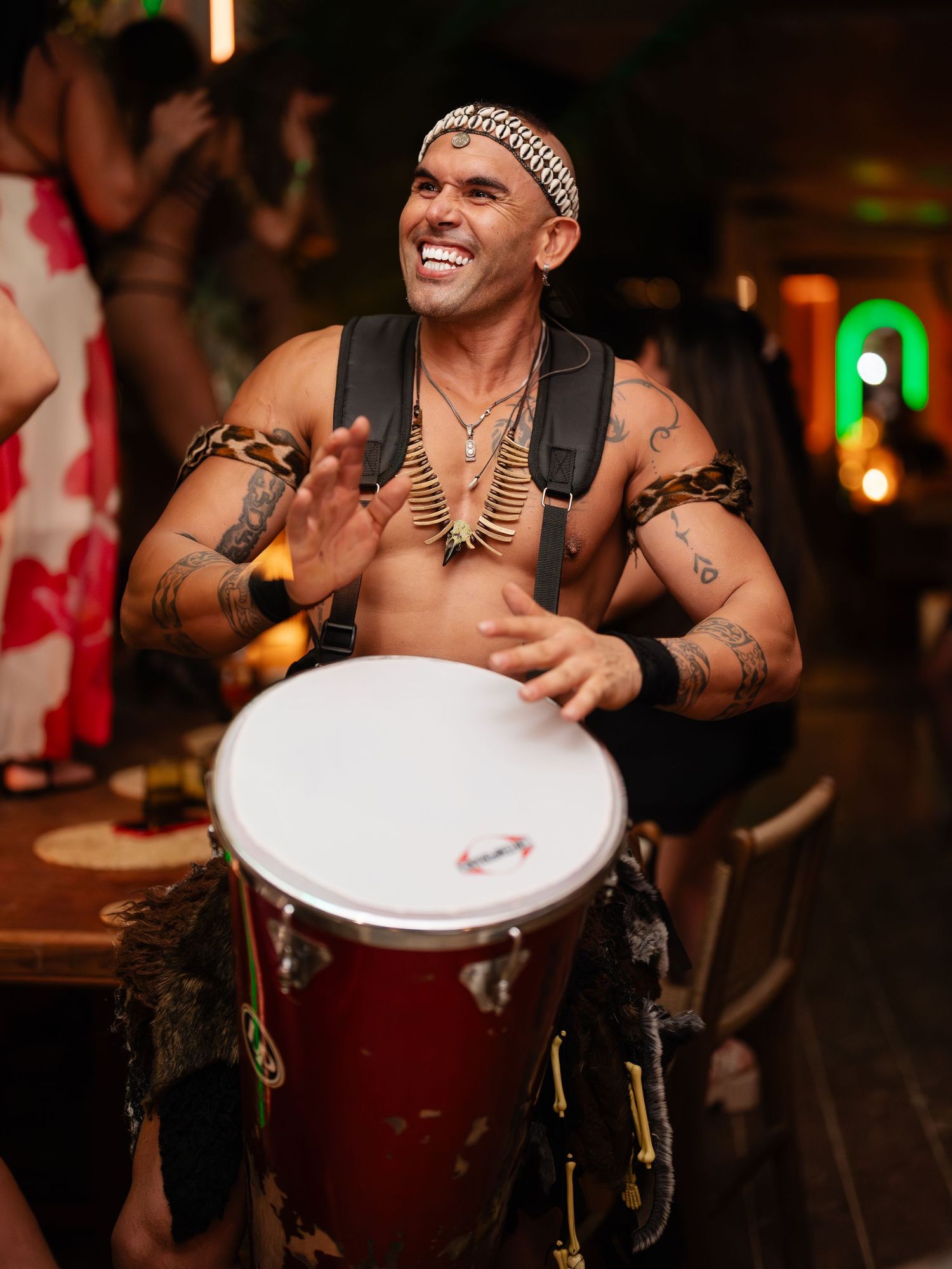 Tattooed male drummer in tribal attire playing a red conga drum at an event.