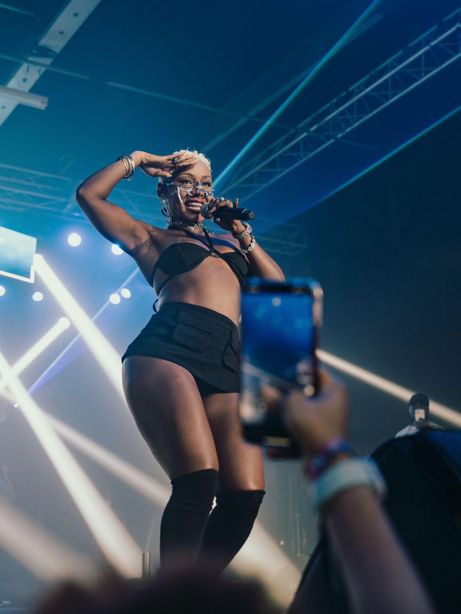 Smiling female performer in black crop top and shorts on stage while fan records with smartphone.