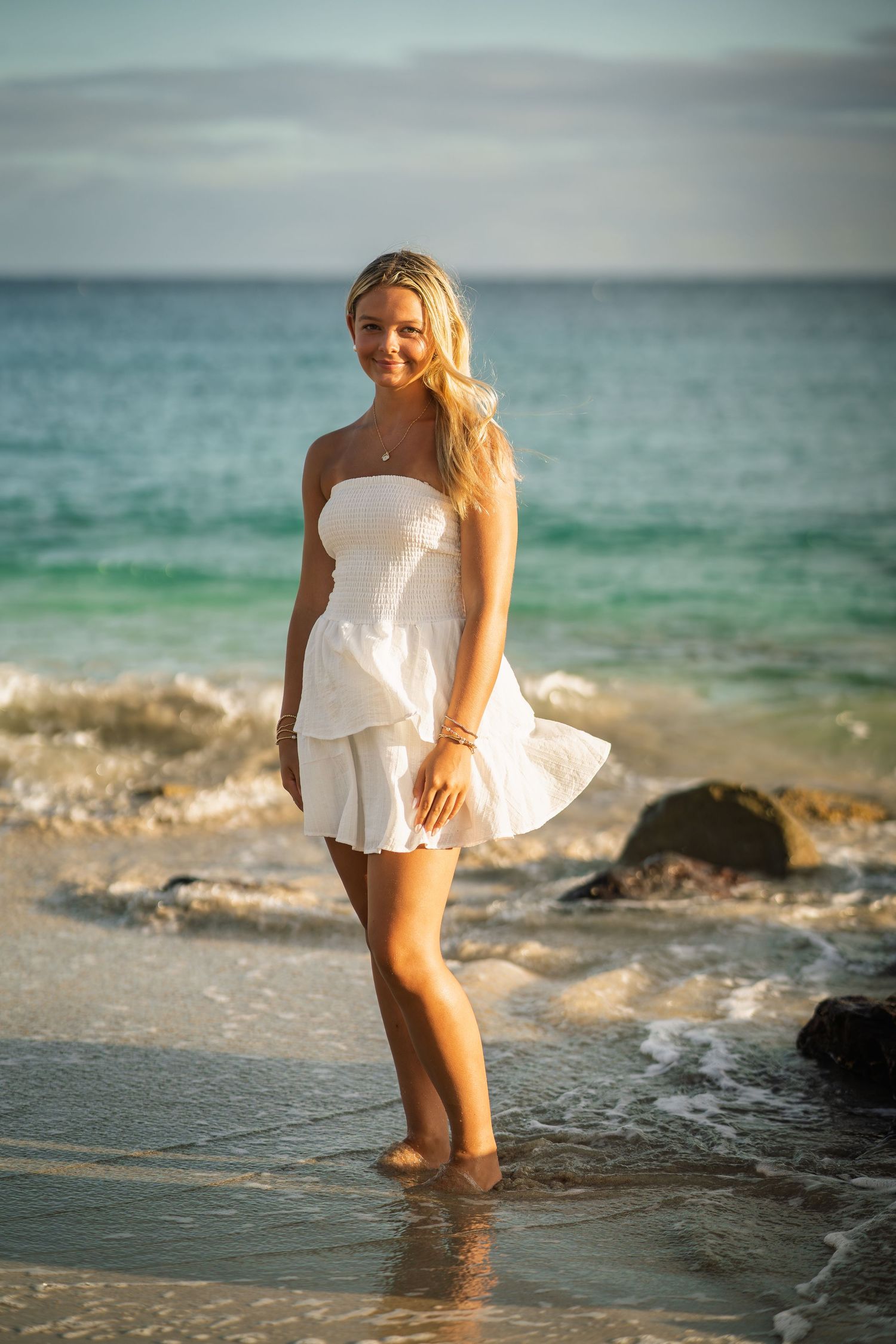 Blonde young woman in white strapless mini dress wading in ocean waves on tropical beach.