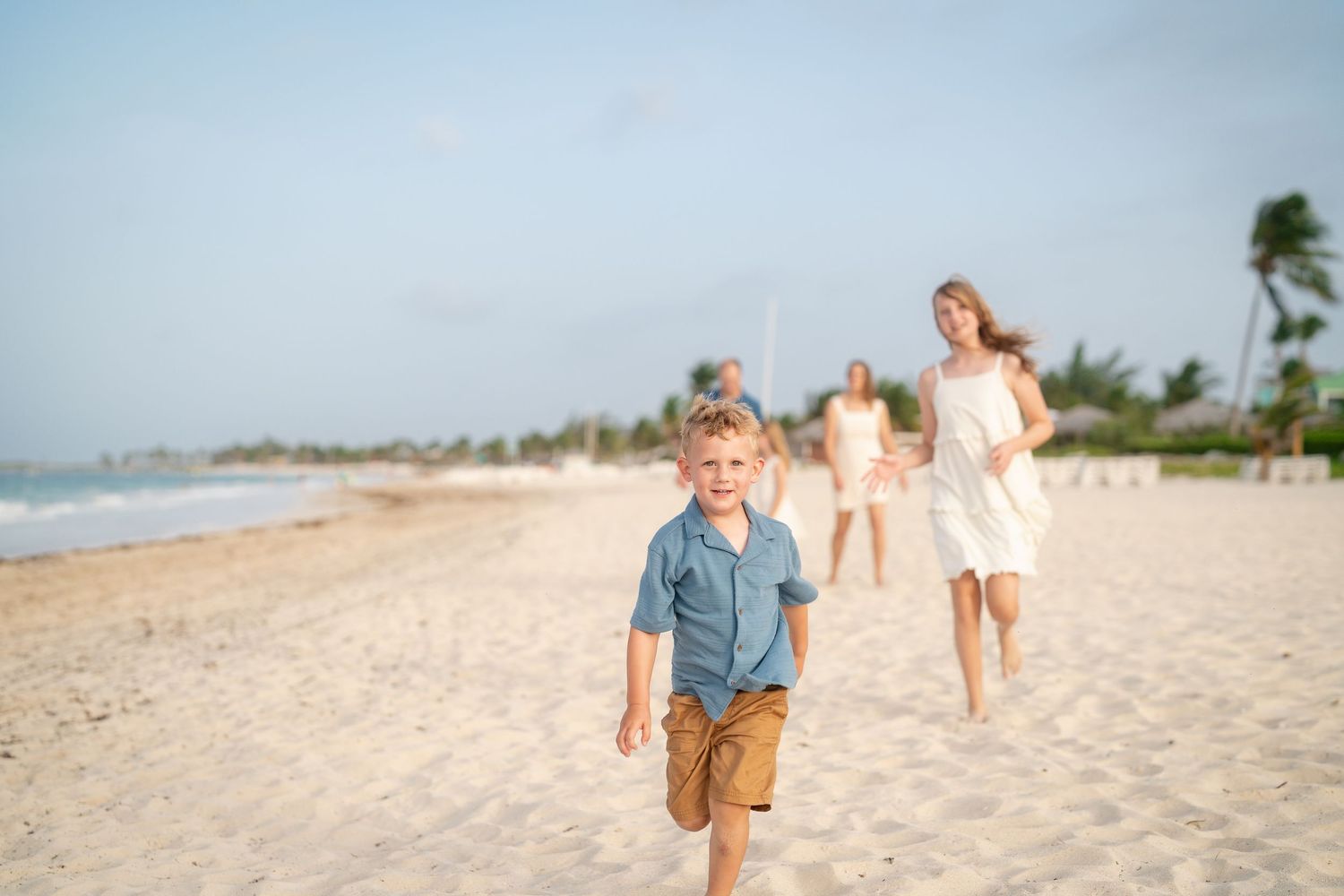 Young blonde boy running on tropical beach with family walking behind him.