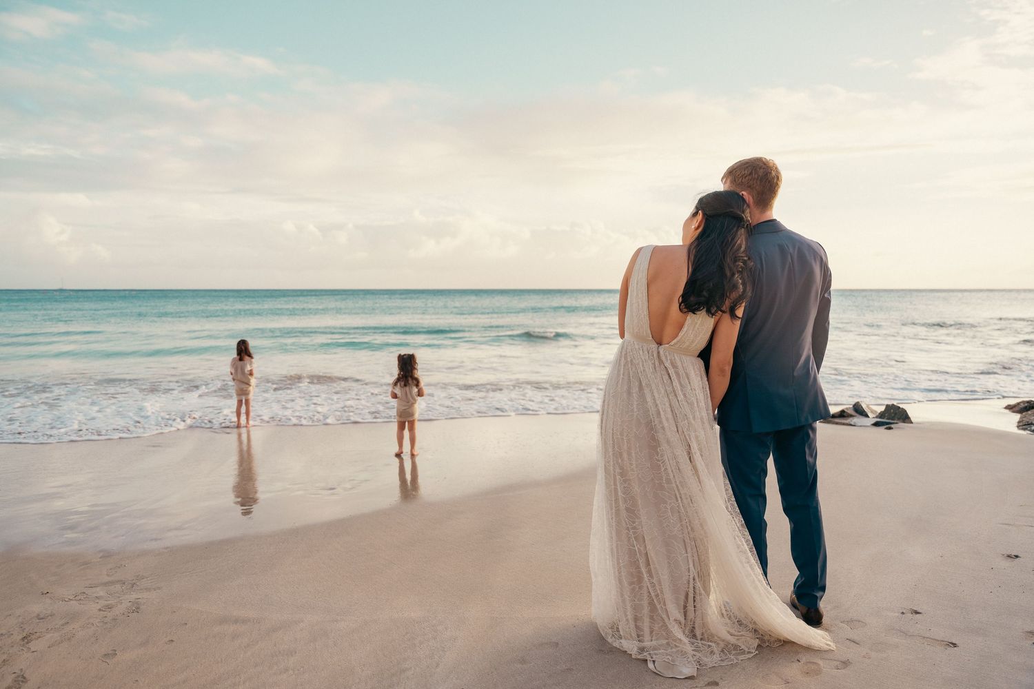 Couple in wedding attire standing on a tropical beach watching two children play at the water's edge at sunset.