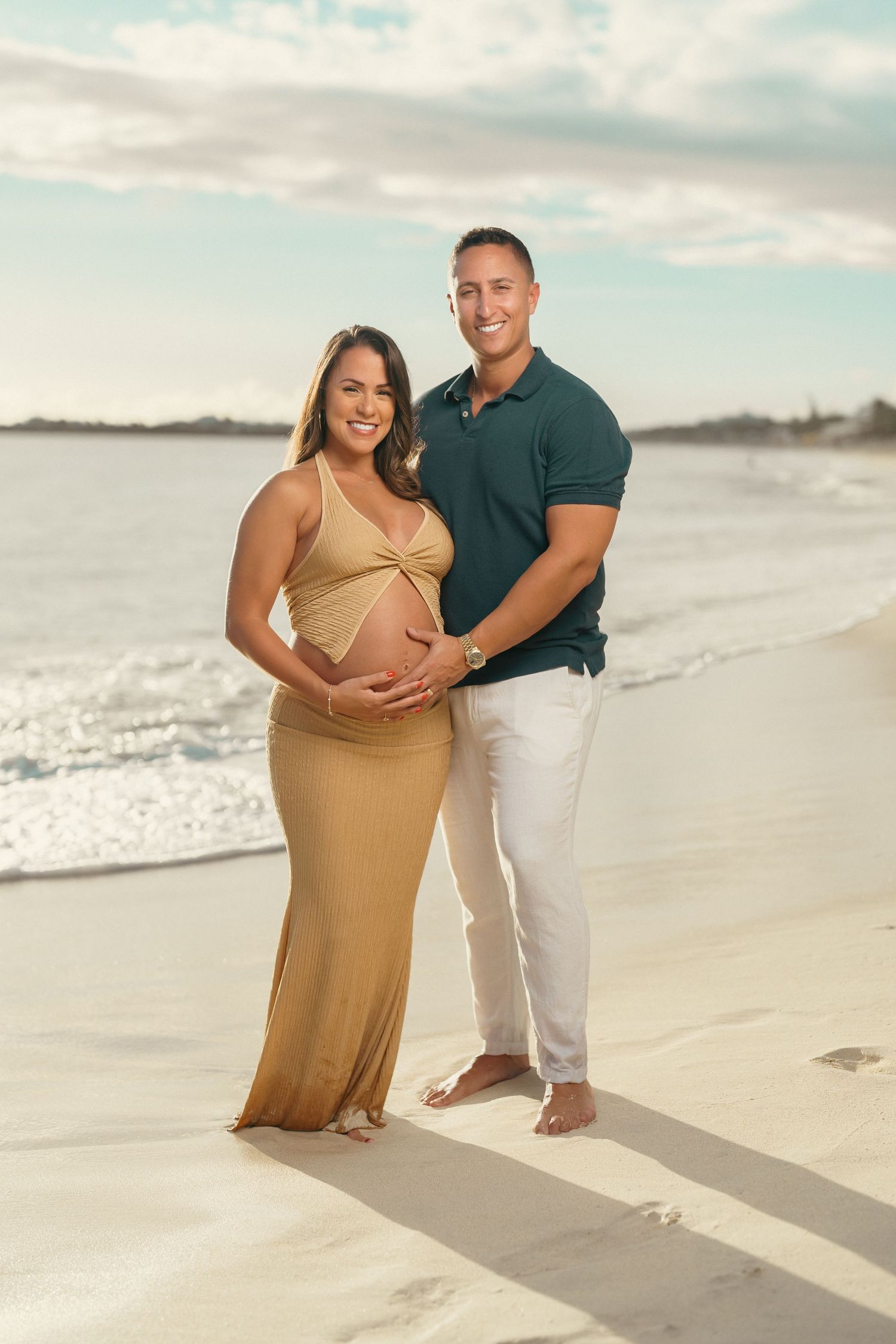Happy couple posing on beach, pregnant woman in gold dress with partner in teal shirt.