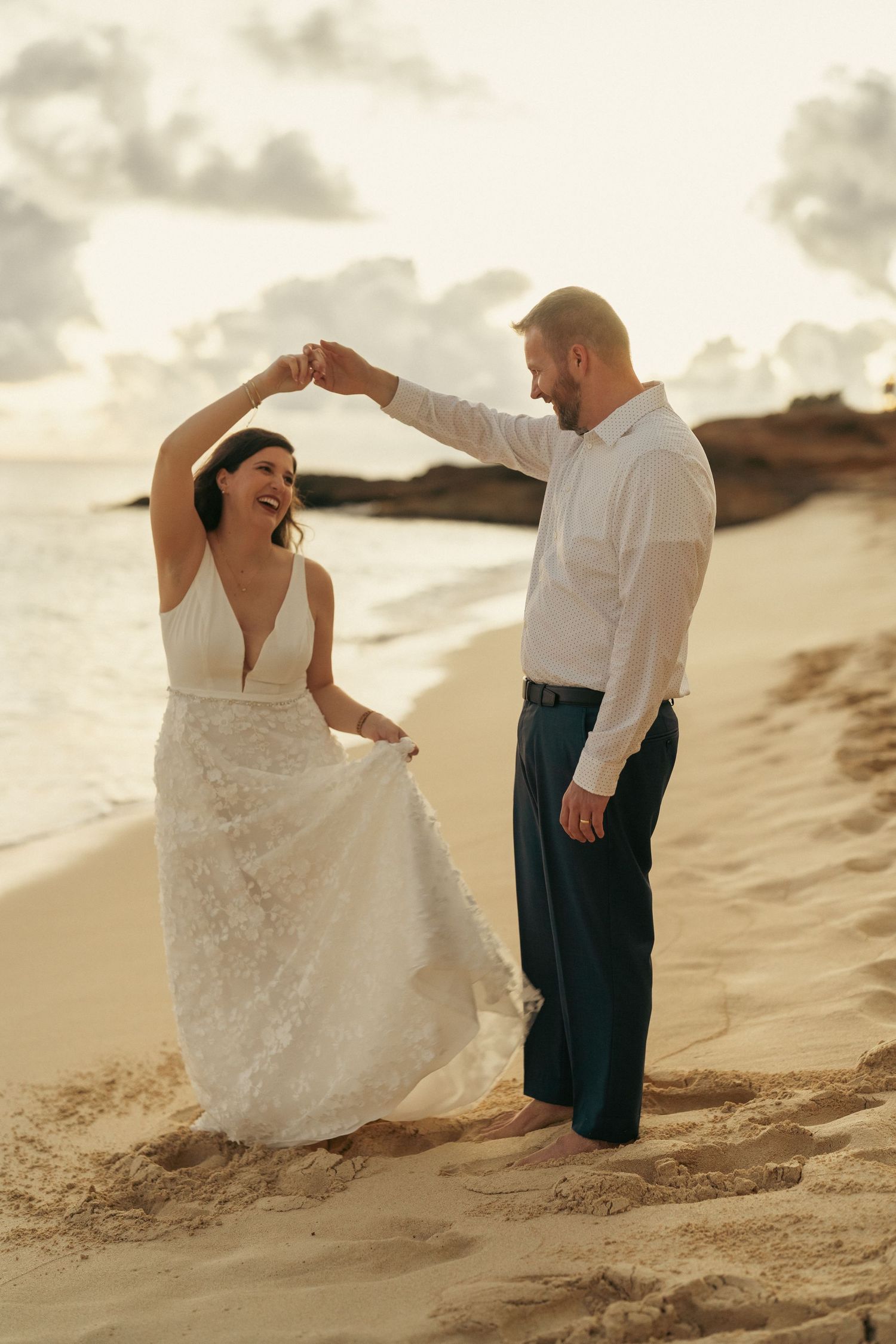 Newlywed couple dancing on a sandy beach at sunset, bride in a lace wedding gown.