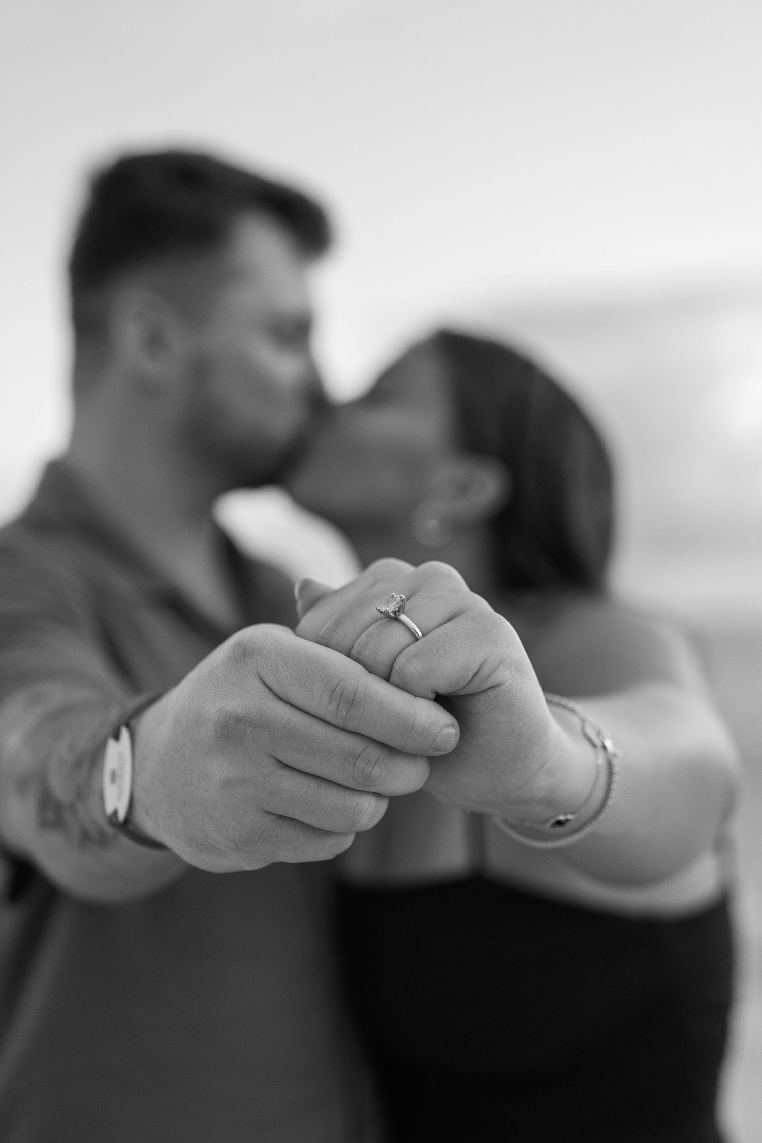 Engaged couple kissing while showing off diamond engagement ring in black and white beach photo.