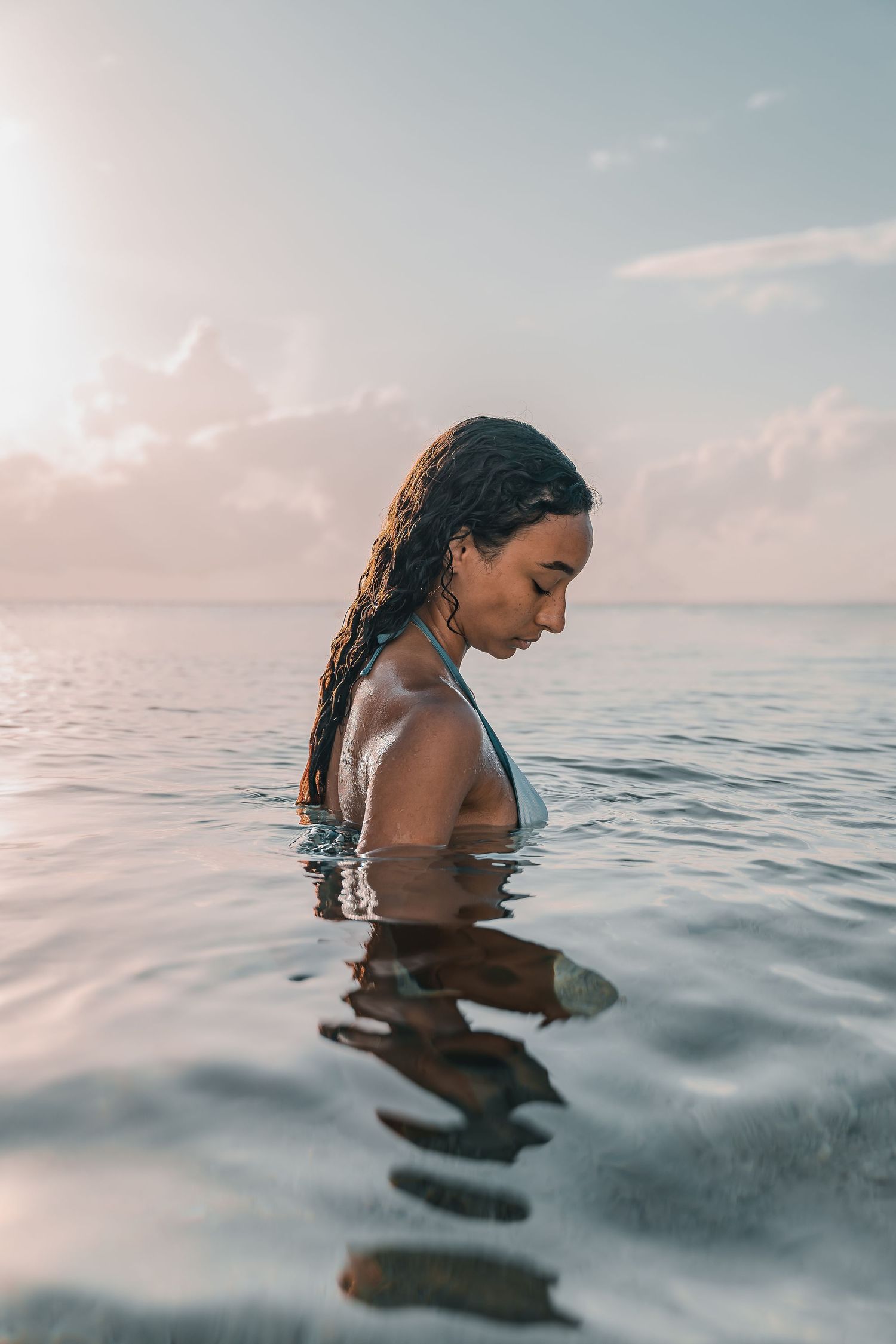 Woman with wet braided hair standing in calm ocean water at sunset, looking down peacefully.