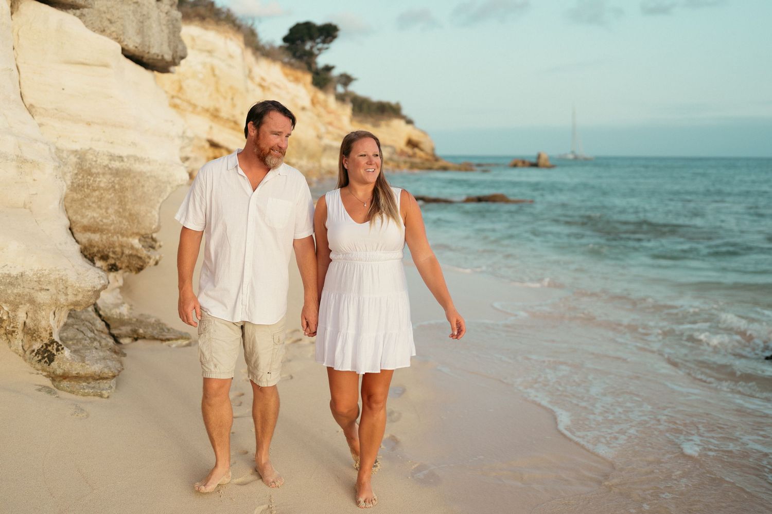 A happy couple in white outfits walks barefoot along a sandy beach with white cliffs at sunset.